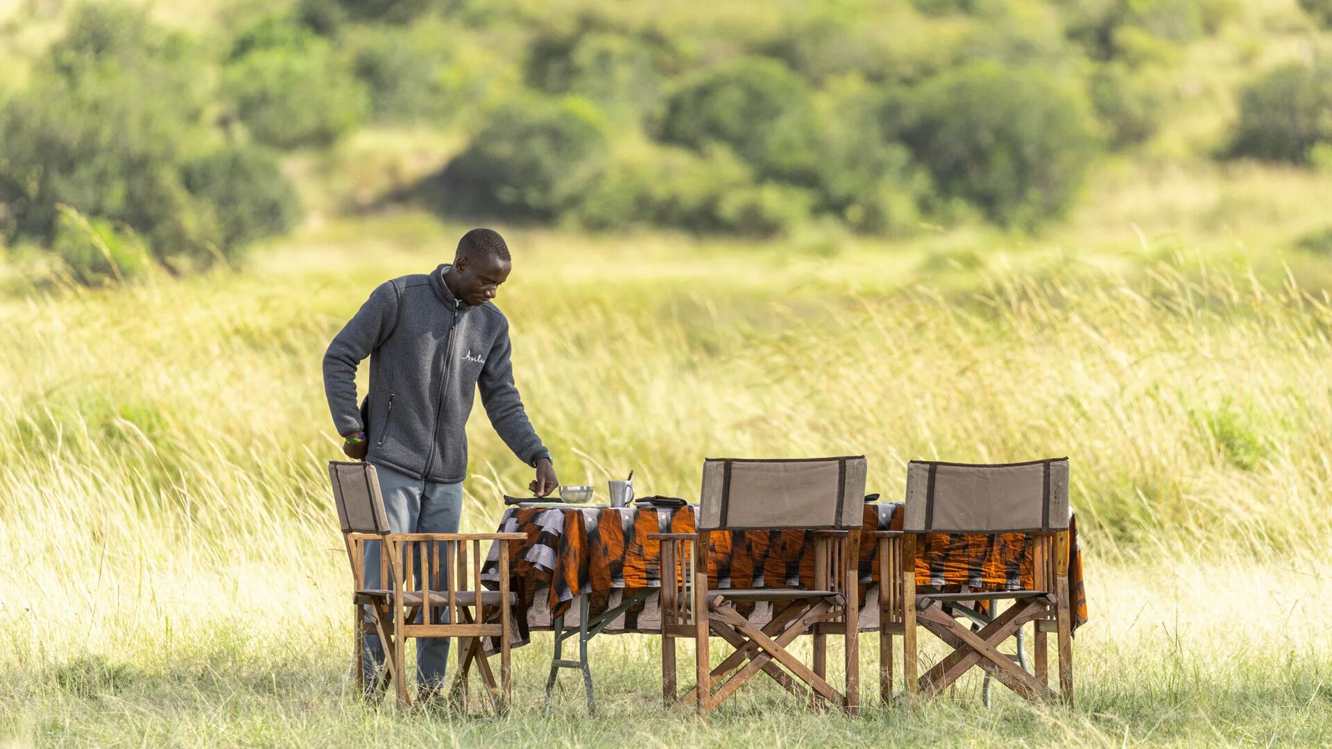 A staff member sets a dining table in tall grass for a bush lunch, with folding chairs arranged in open savannah and green woodland in the distance.