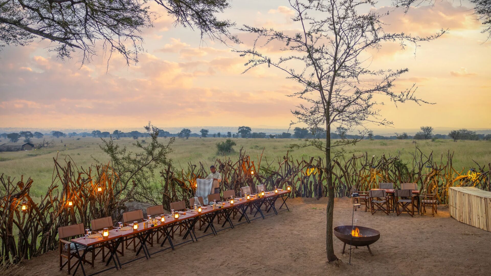 An outdoor safari dinner set at dusk, with long tables lit by lanterns, a small fire pit, rustic fencing, and expansive savannah views beneath a soft sunset sky.