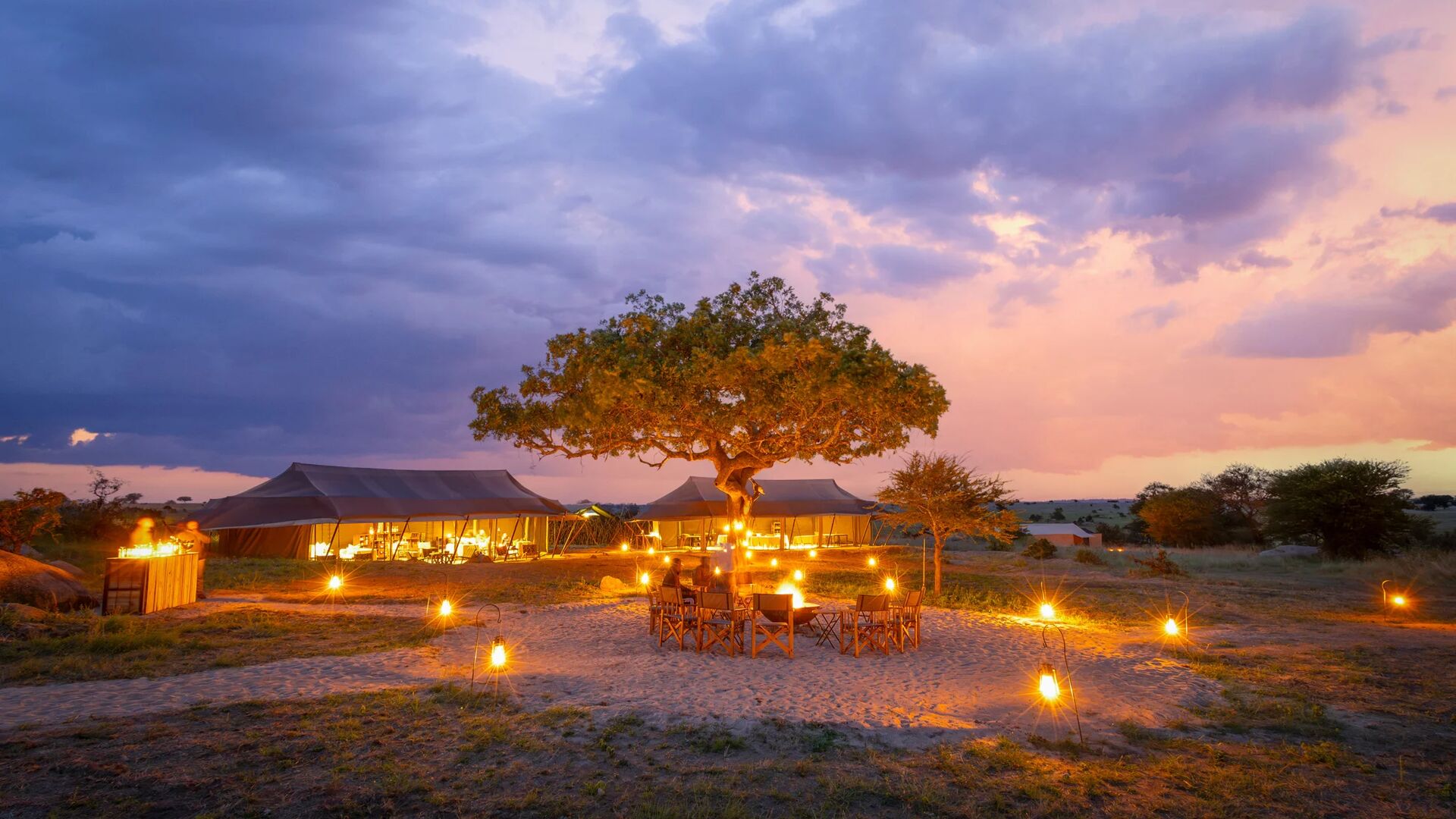 Olakira Migration Camp glows at night with lanterns surrounding outdoor seating beneath a large tree, canvas tents illuminated in the background under colourful dusk clouds.