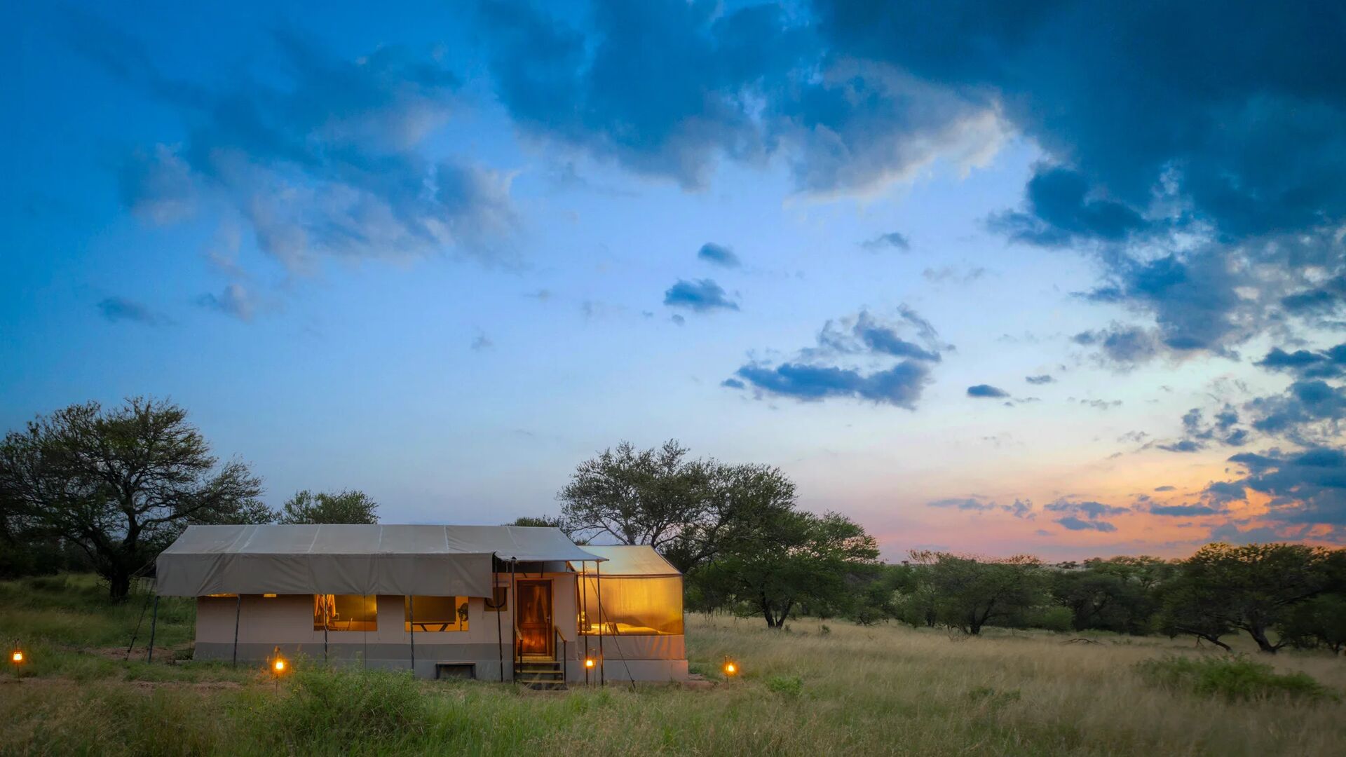 A safari tent softly lit at dusk, set among grassy plains and scattered trees, with lanterns marking the path and a dramatic blue and pink evening sky overhead.