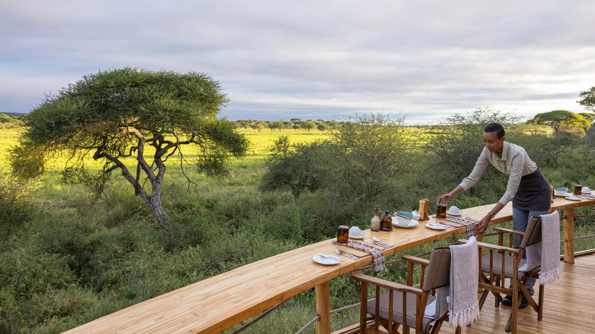 An Asilia staff member sets a breakfast table on a raised wooden deck, with chairs arranged along the edge and expansive views of Tarangire National Park and acacia trees beyond.