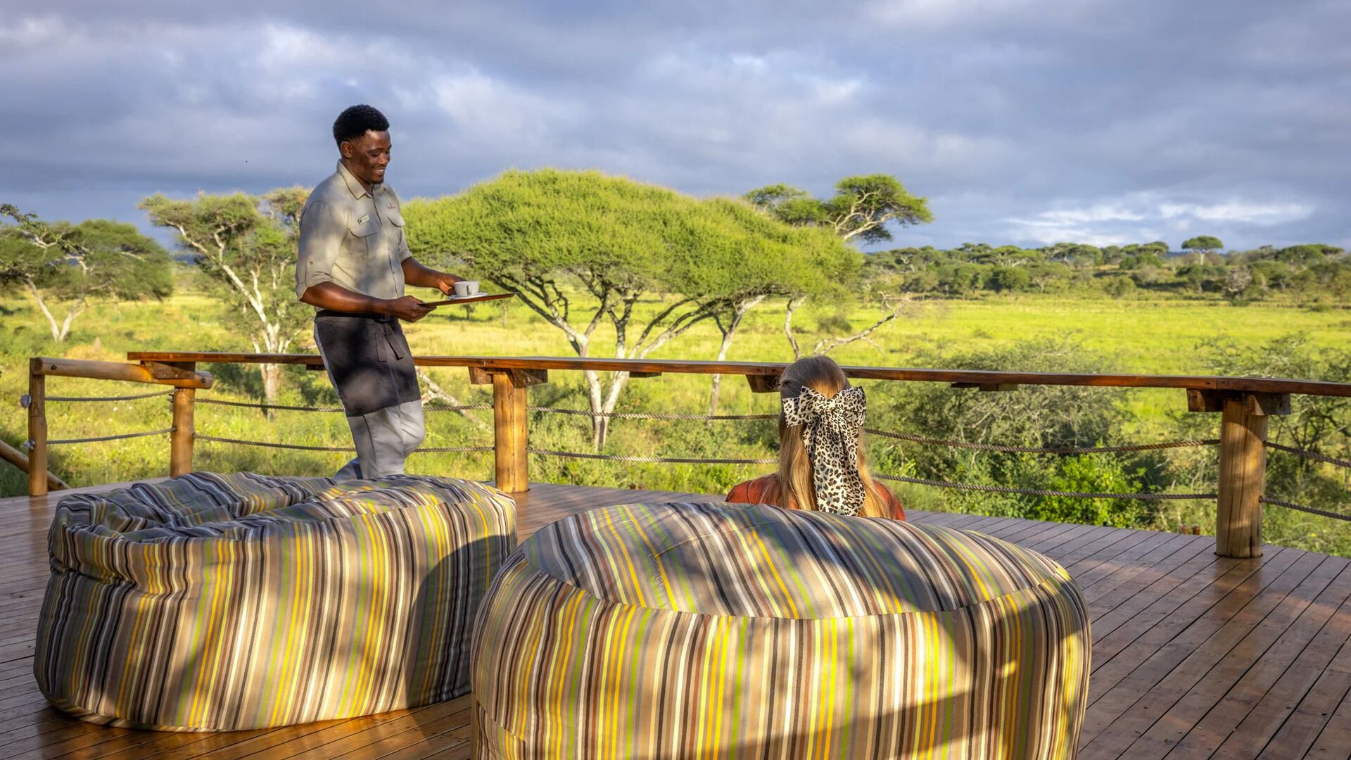 An Asilia staff member serves a drink to a female guest relaxing on a wooden deck, with comfortable seating and sweeping views of Tarangire National Park.