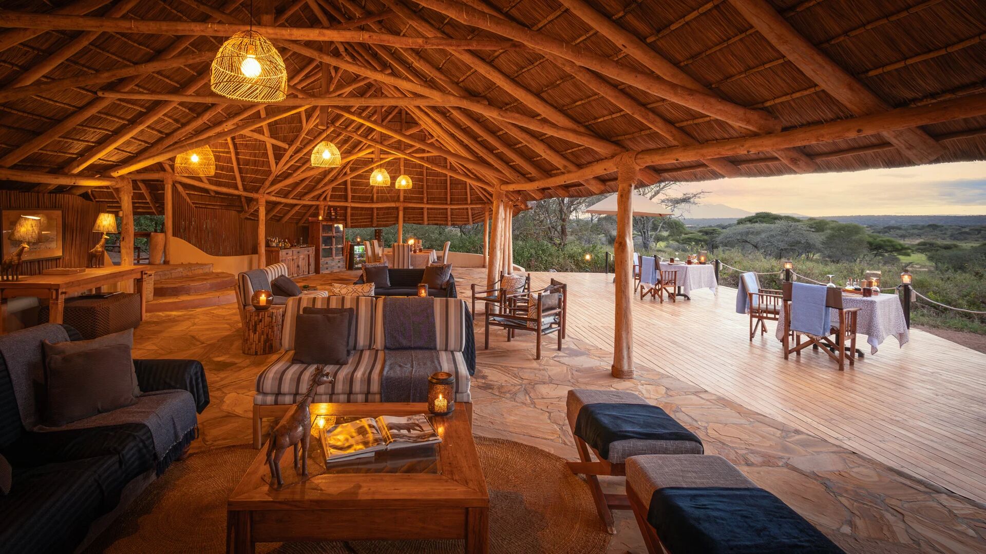 The spacious open-sided lounge and dining area at Oliver's Camp with warm lantern lighting, wooden beams, and expansive views across surrounding bushland at dusk.