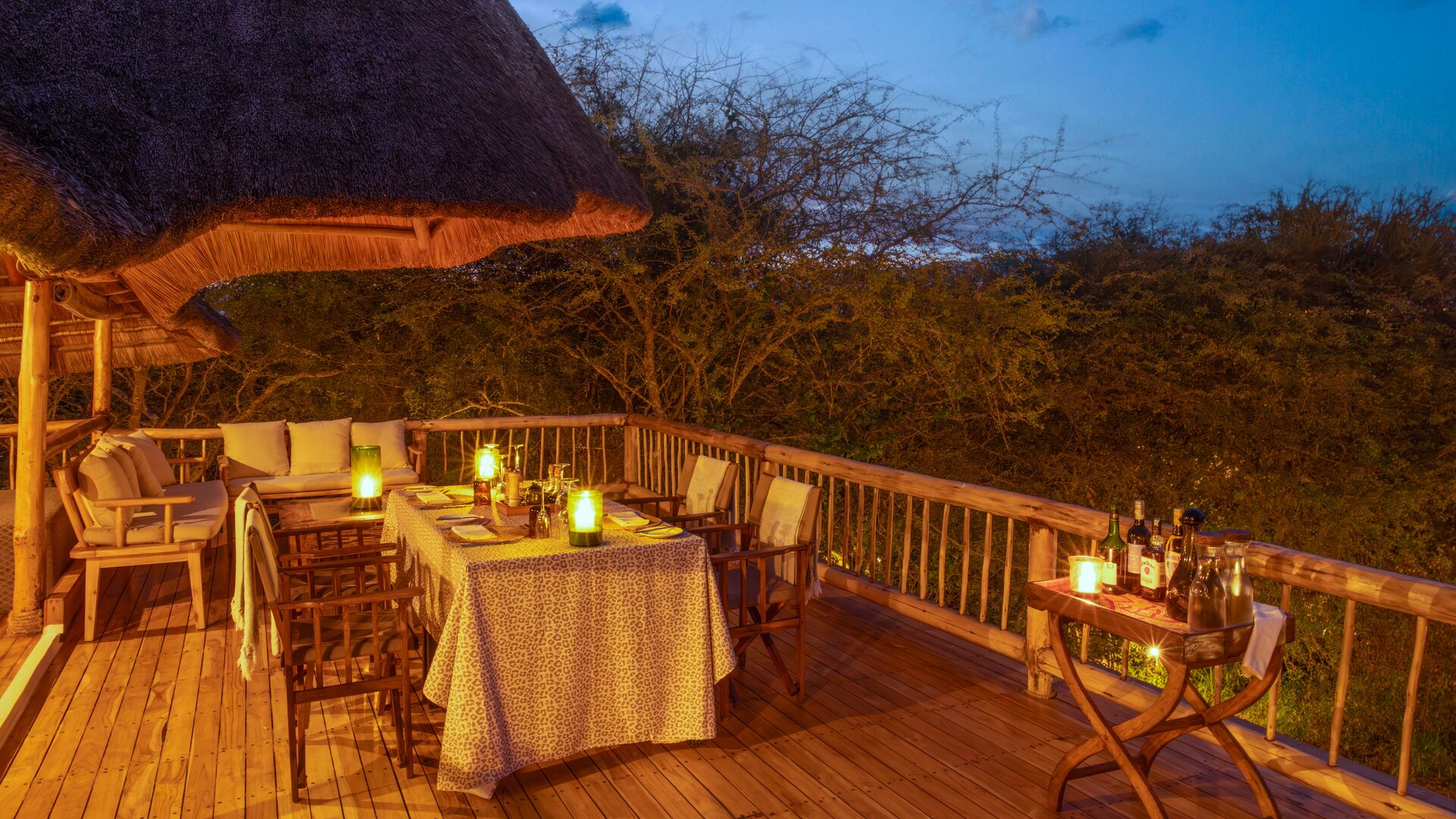 A wooden deck dining area with a table set for a meal, surrounded by lanterns and overlooking the bush at dusk