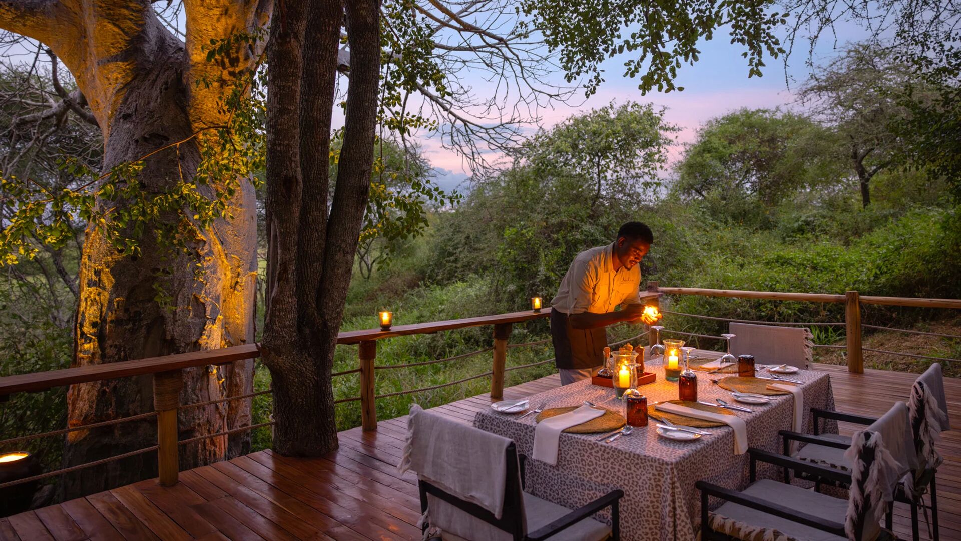 A staff member sets a candlelit dining table at Oliver's Camp on a wooden deck surrounded by trees, creating an intimate outdoor dinner setting at dusk.