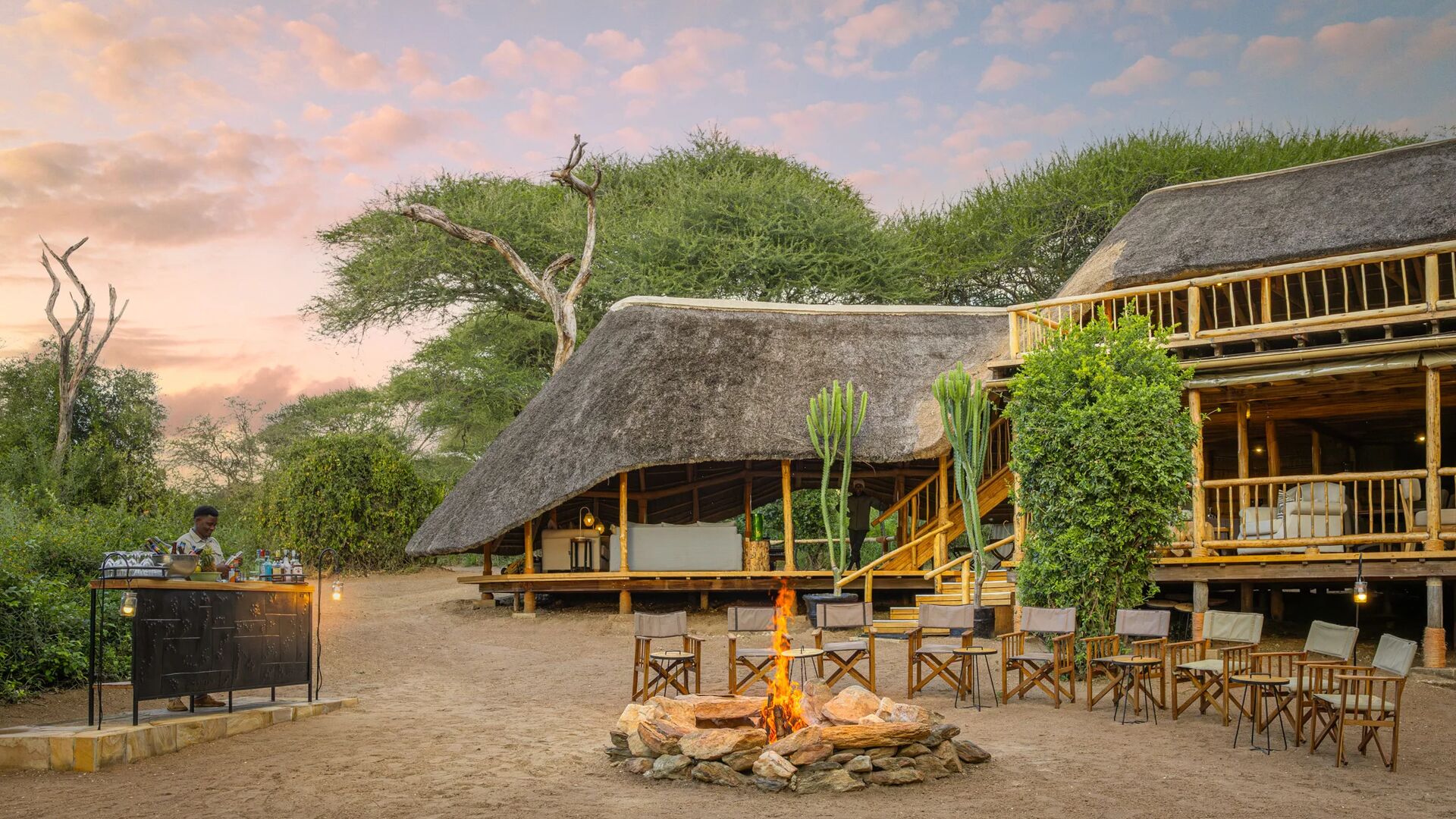 Outdoor area at Oliver's Camp with thatched buildings, a central fire pit surrounded by chairs, and a bar setup, set among trees at dusk with warm lantern light.