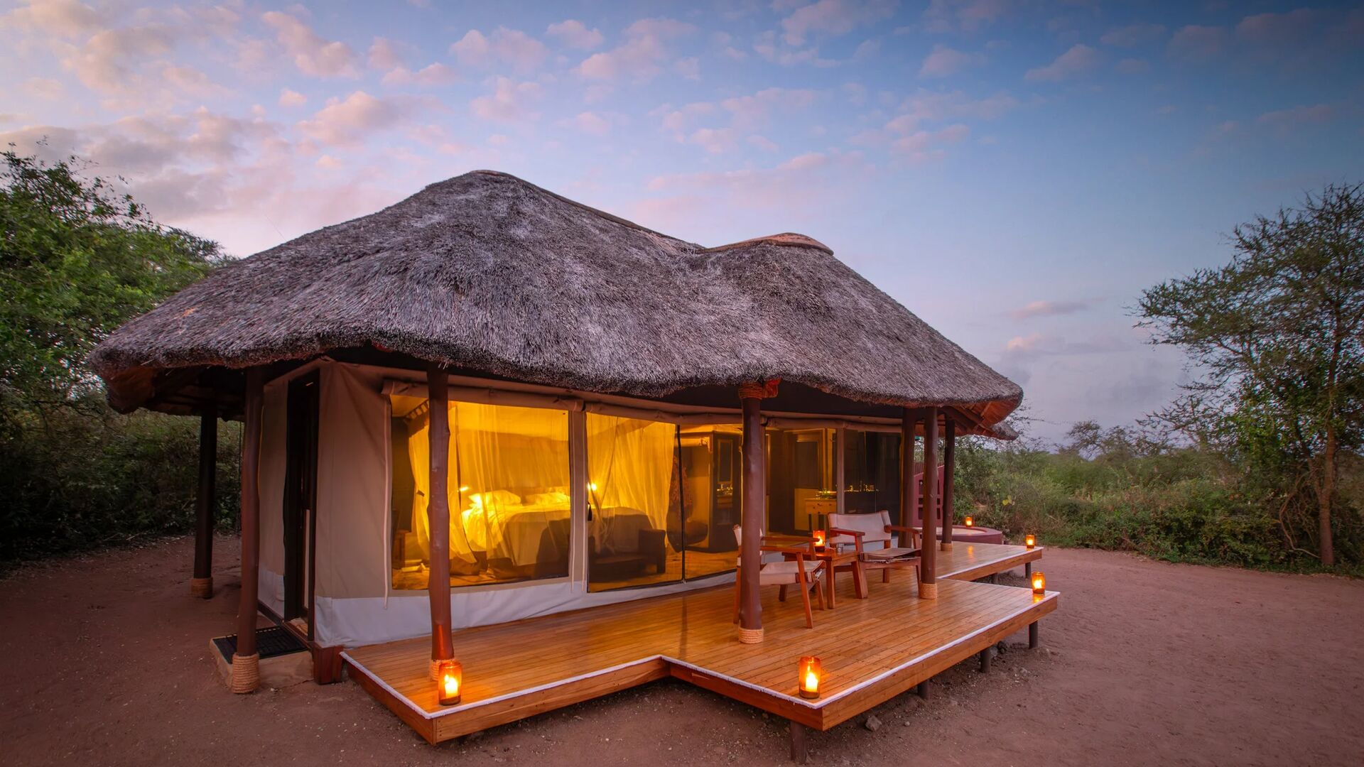 The exterior of a luxury safari tent at Oliver's Camp with a thatched roof, wooden deck, and lanterns, softly lit at dusk and surrounded by natural bushland.