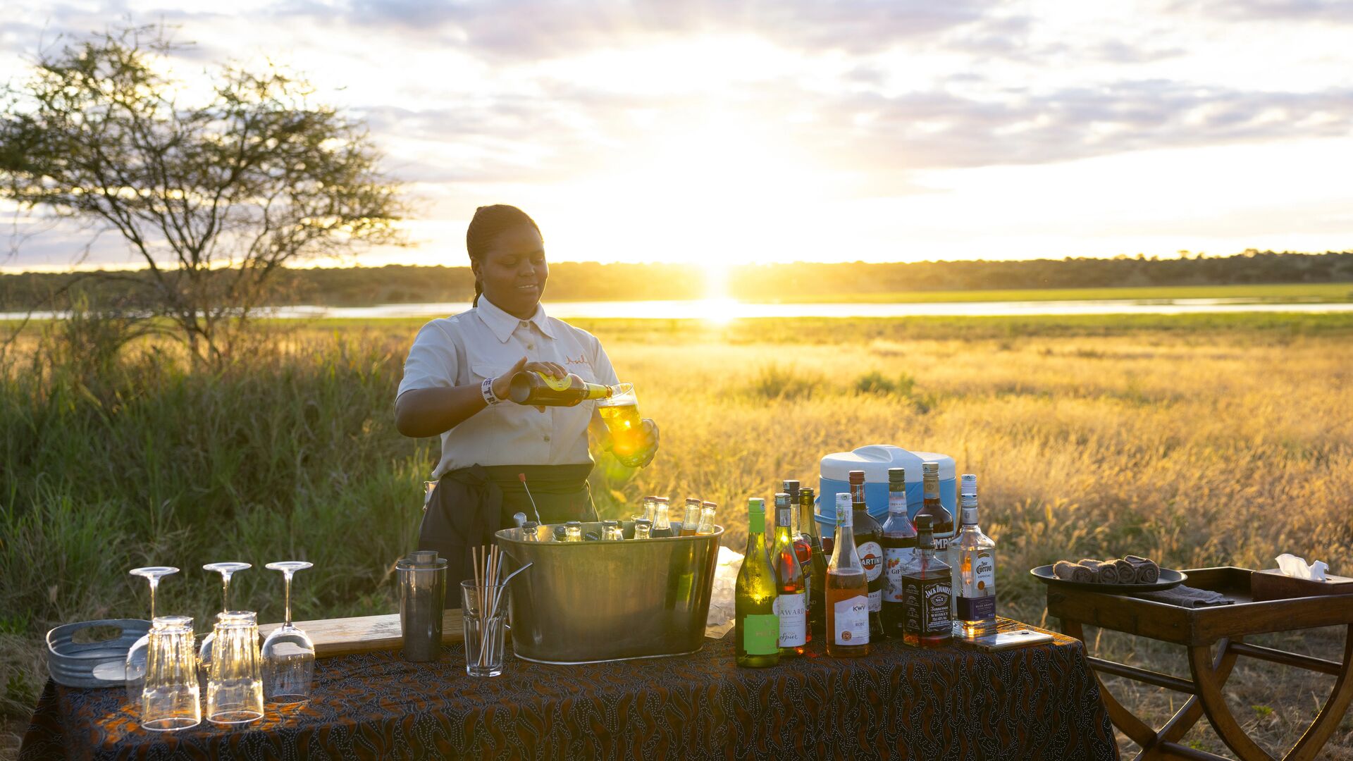 An Asilia staff member pours a drink at a bush bar setup with bottles and glassware during sunset