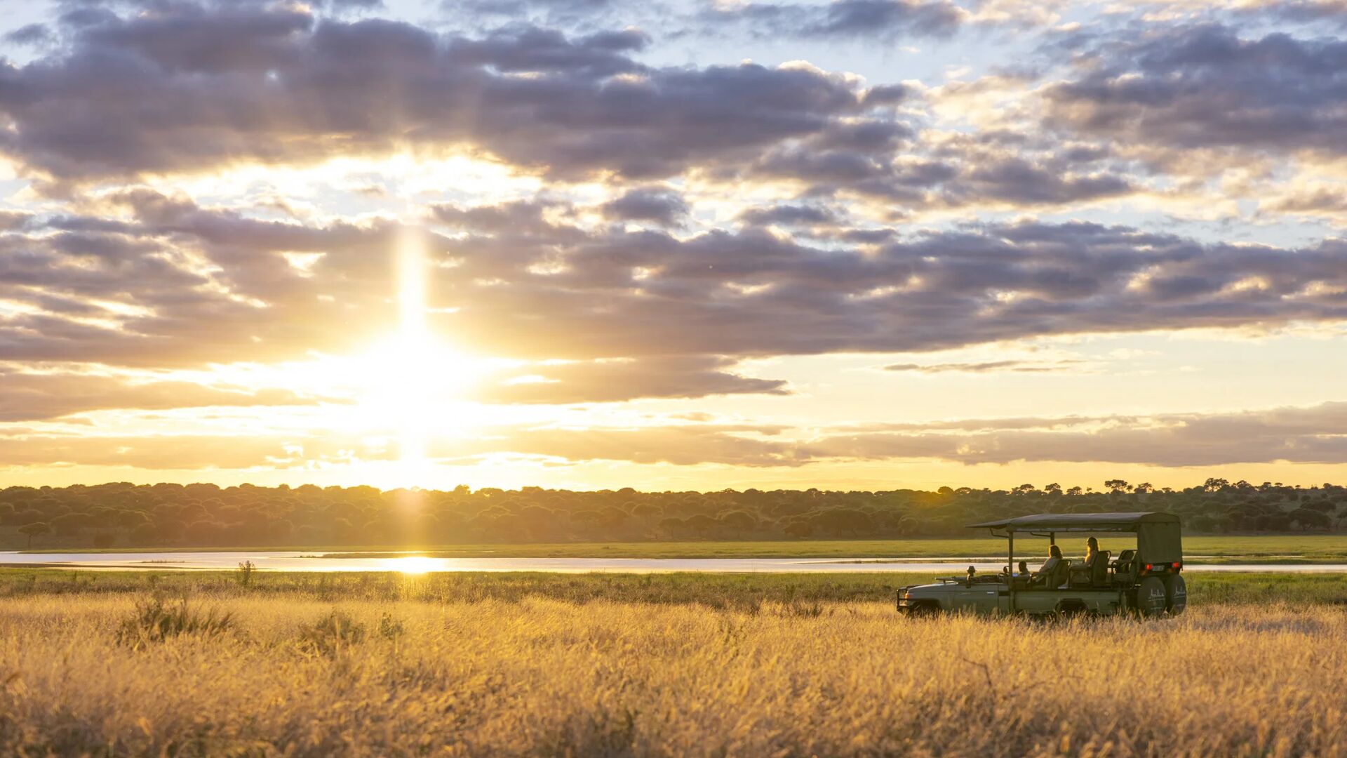 An Asilia safari vehicle pauses in golden grass at sunset, with guests watching the glowing sky reflected on a waterhole, framed by distant trees and layered clouds.