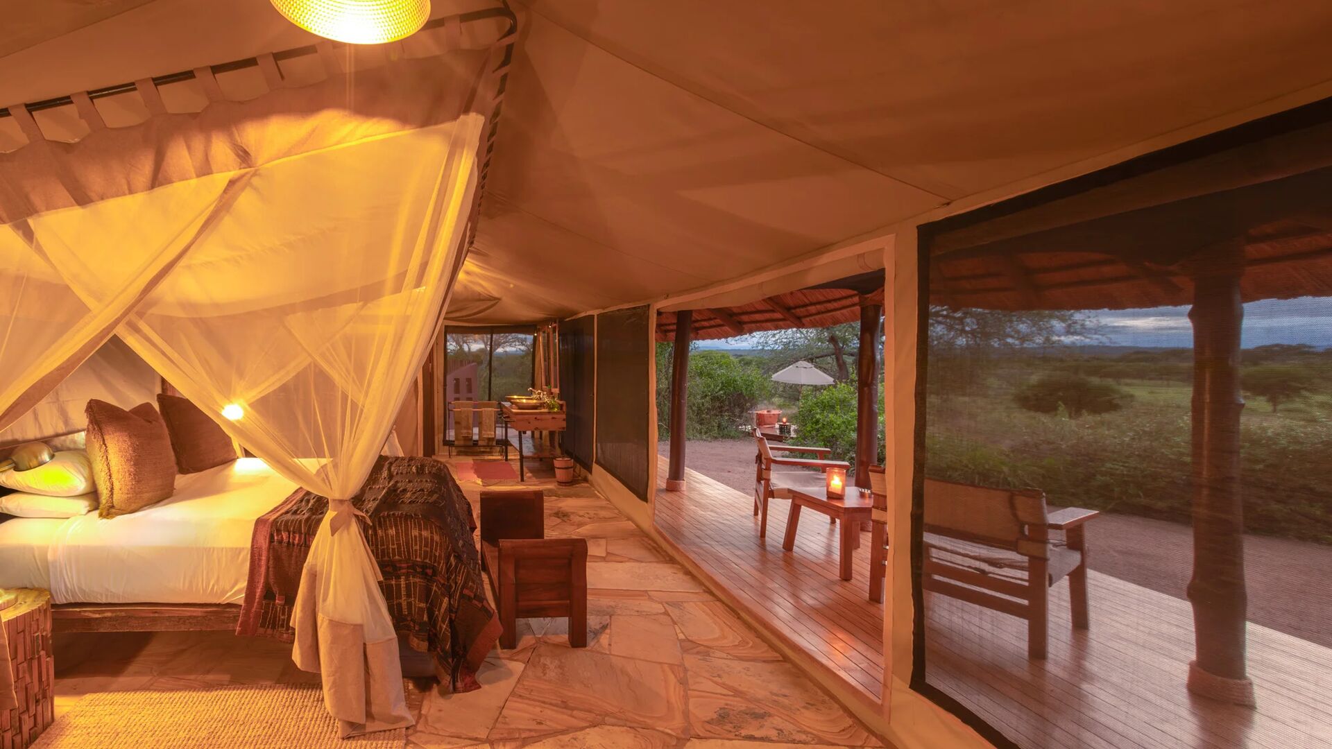 The interior of a luxury safari tent at Oliver's Camp with a draped mosquito-net bed, warm lighting, and stone floors, opening onto a private wooden deck overlooking bushland at dusk.