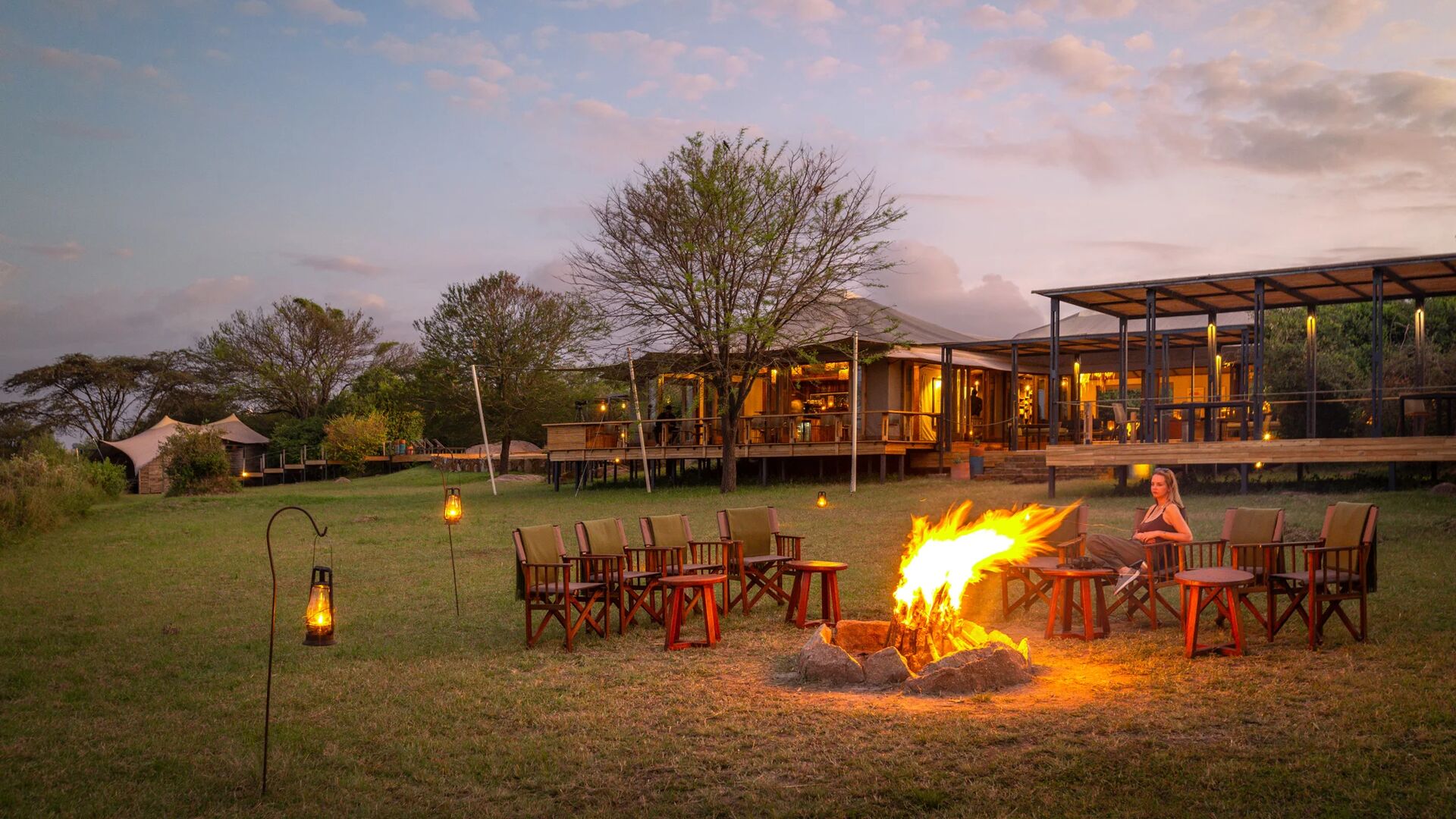 Guests relax around a glowing campfire at dusk, with wooden safari chairs arranged on the grass and the warmly lit Sayari Camp lodge in the background under a soft pink and blue evening sky.