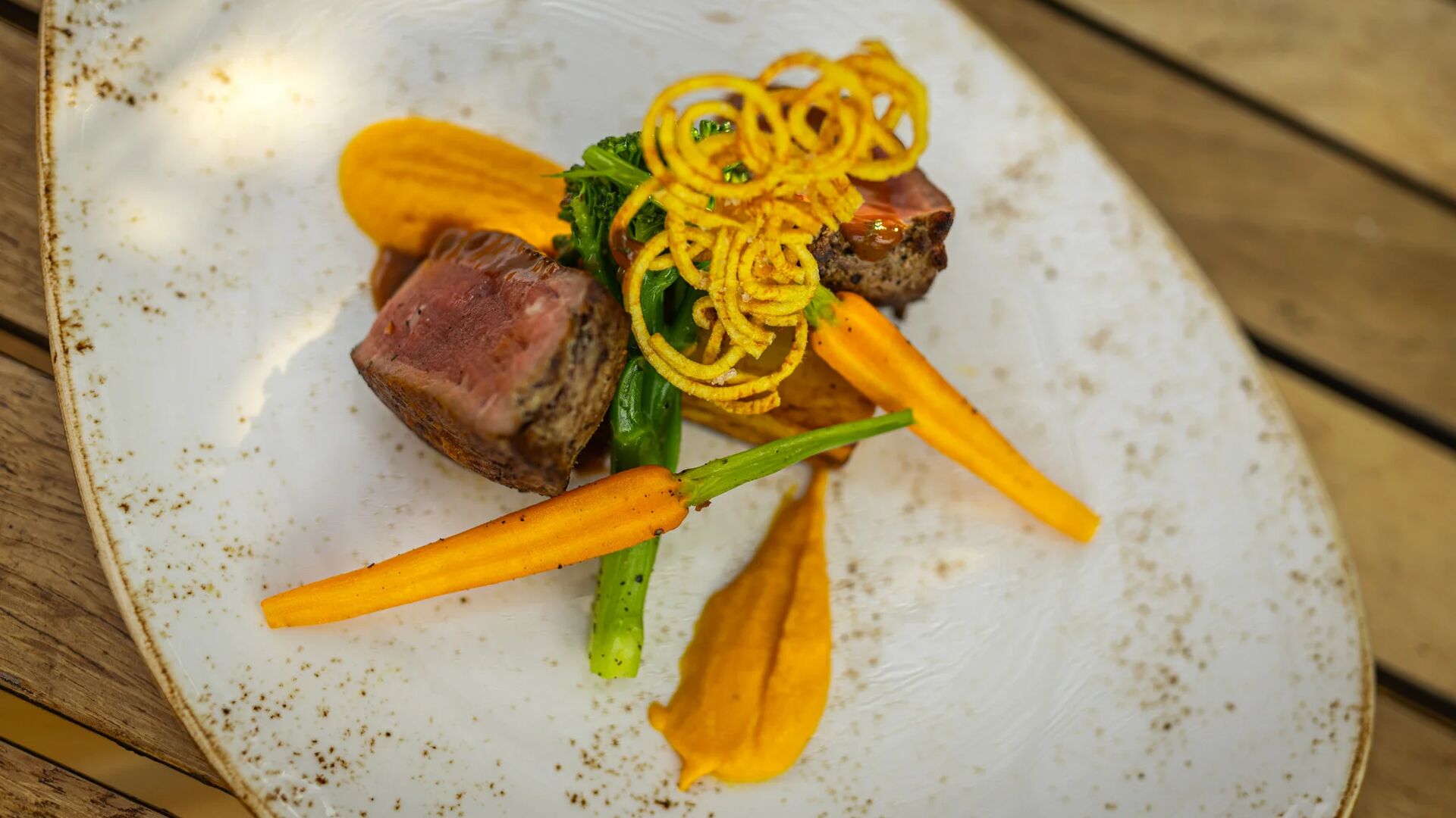 An elegant fine-dining plate featuring sliced, medium-rare beef served with tender green vegetables, roasted baby carrots, smooth carrot purée, and crisp golden garnish, presented on a rustic ceramic plate atop a wooden table.