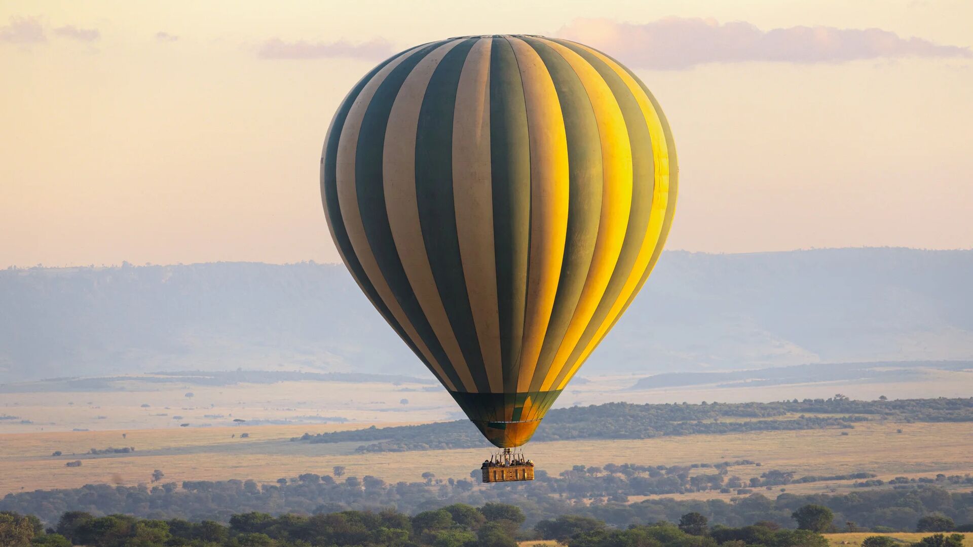 A large striped hot air balloon floats above the Serengeti plains at sunrise, carrying guests in a basket as golden light illuminates rolling grasslands, scattered trees, and distant hills.