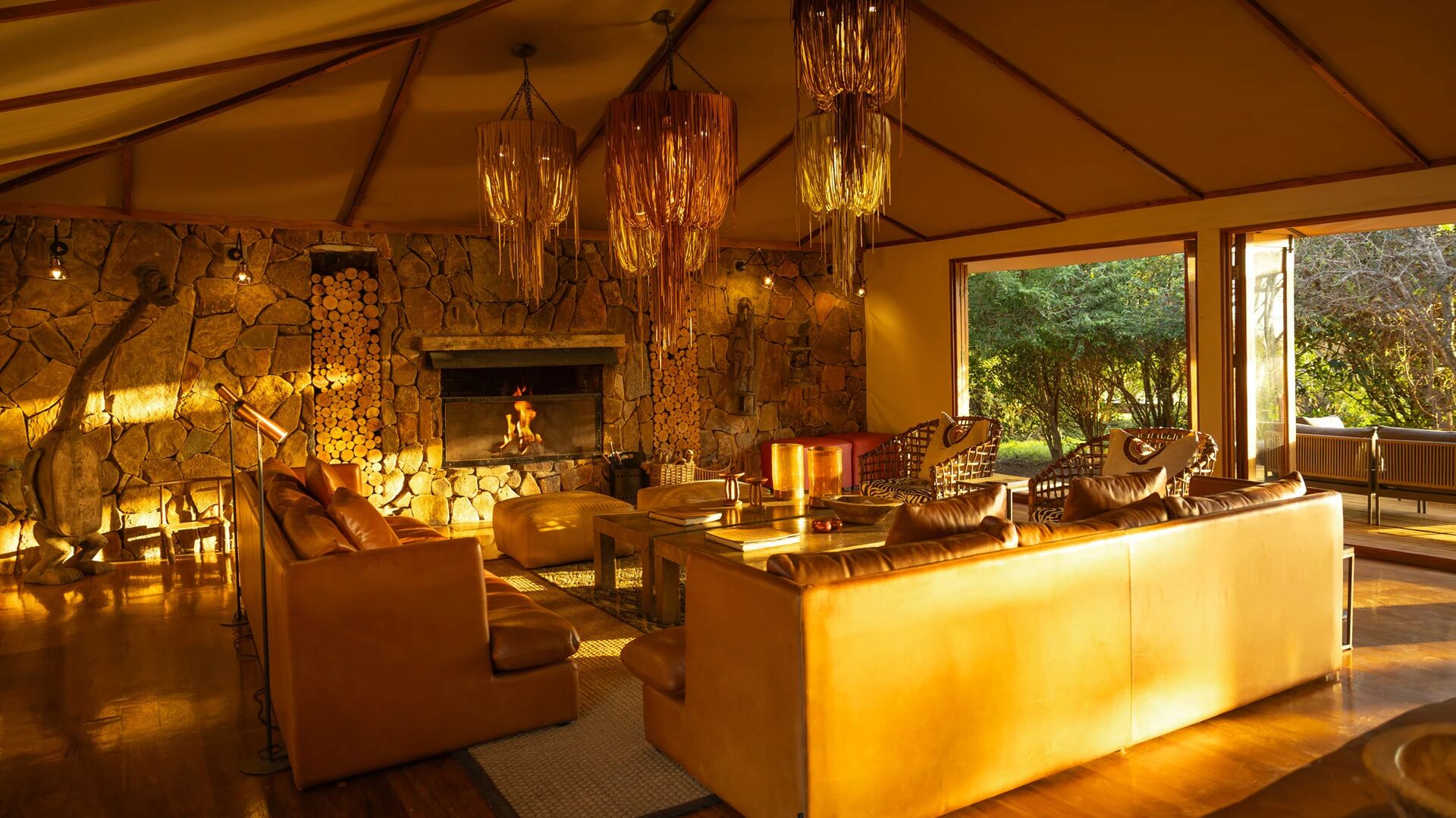 Sayari Camp's cosy lounge with leather sofas, a stone fireplace glowing with firelight, stacked firewood, woven pendant lights, and large open doors looking out onto green woodland.
