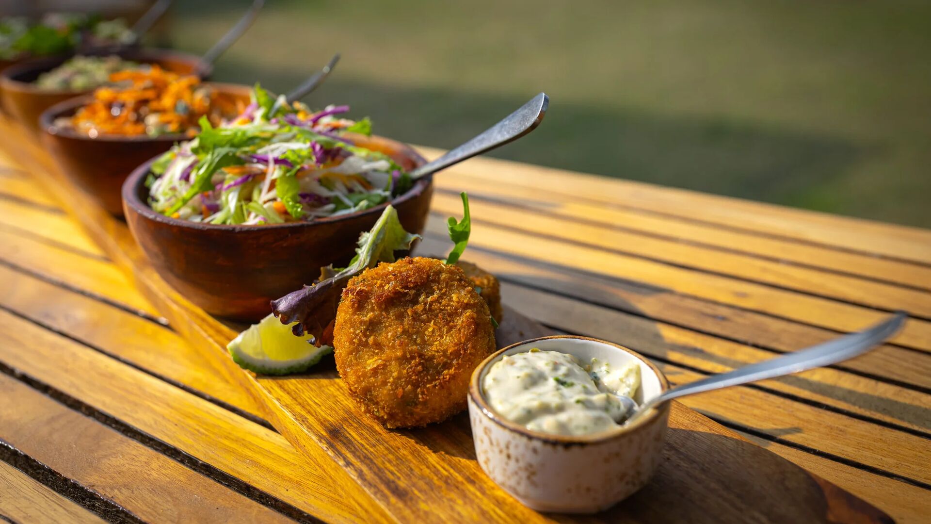 Close-up of a plated lunch at Sayari Camp on a wooden table, featuring golden croquettes with dipping sauce, fresh mixed salads in wooden bowls, and vibrant vegetables, set outdoors in warm natural light.
