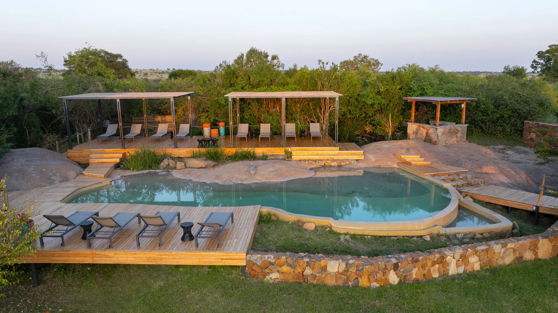 Sayari Camp’s natural stone and wood swimming pool surrounded by sun loungers and shaded decks, set among green trees and open Serengeti plains in soft morning light.