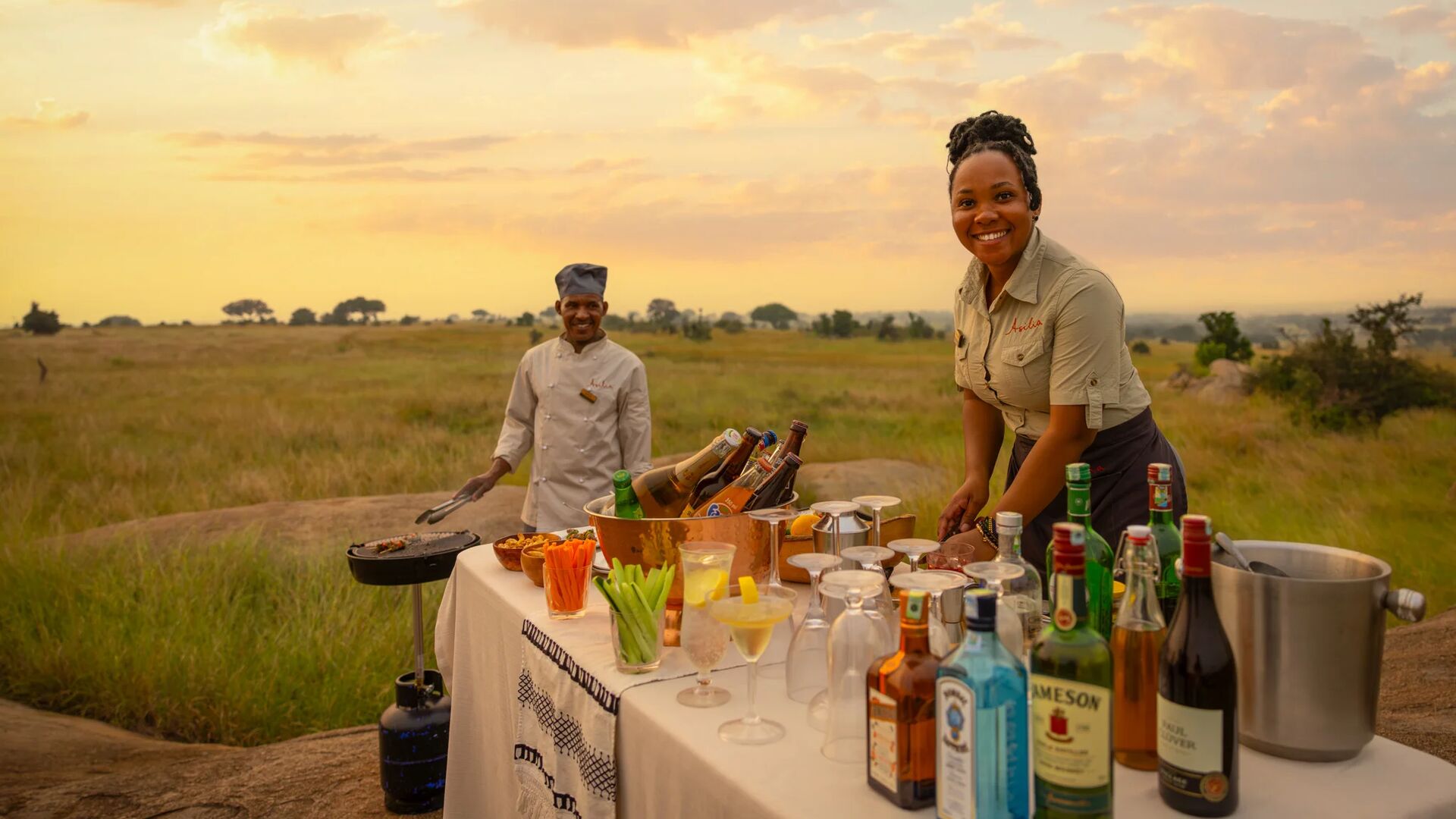 Two Sayari Camp staff prepare a classic safari sundowner setup on the open plains, with a table of drinks, glasses, and snacks at sunset, golden grasslands stretching into the distance beneath a warm evening sky.