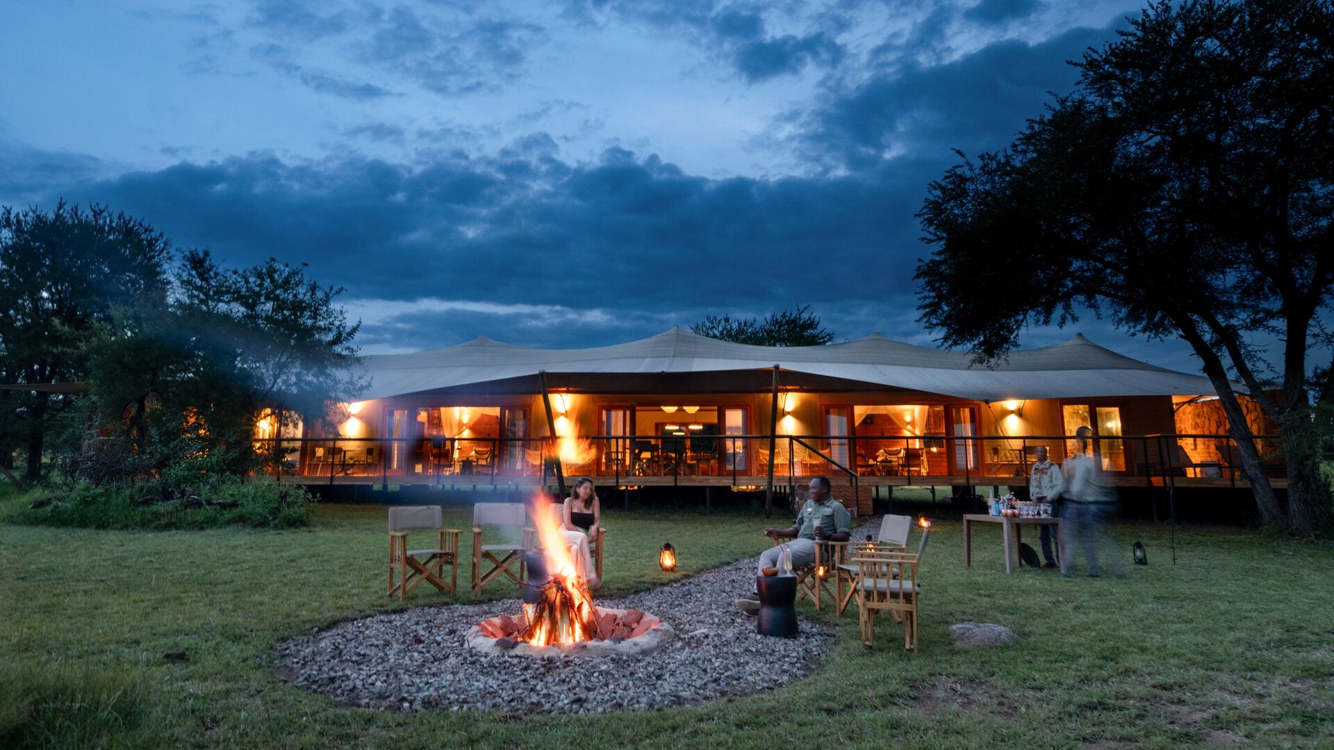 Guests sit around a glowing campfire on a grassy lawn at dusk, with lanterns and chairs arranged in a circle, while the warmly lit Sayari Retreat lodge stands behind them under a deep blue, cloud-filled evening sky.