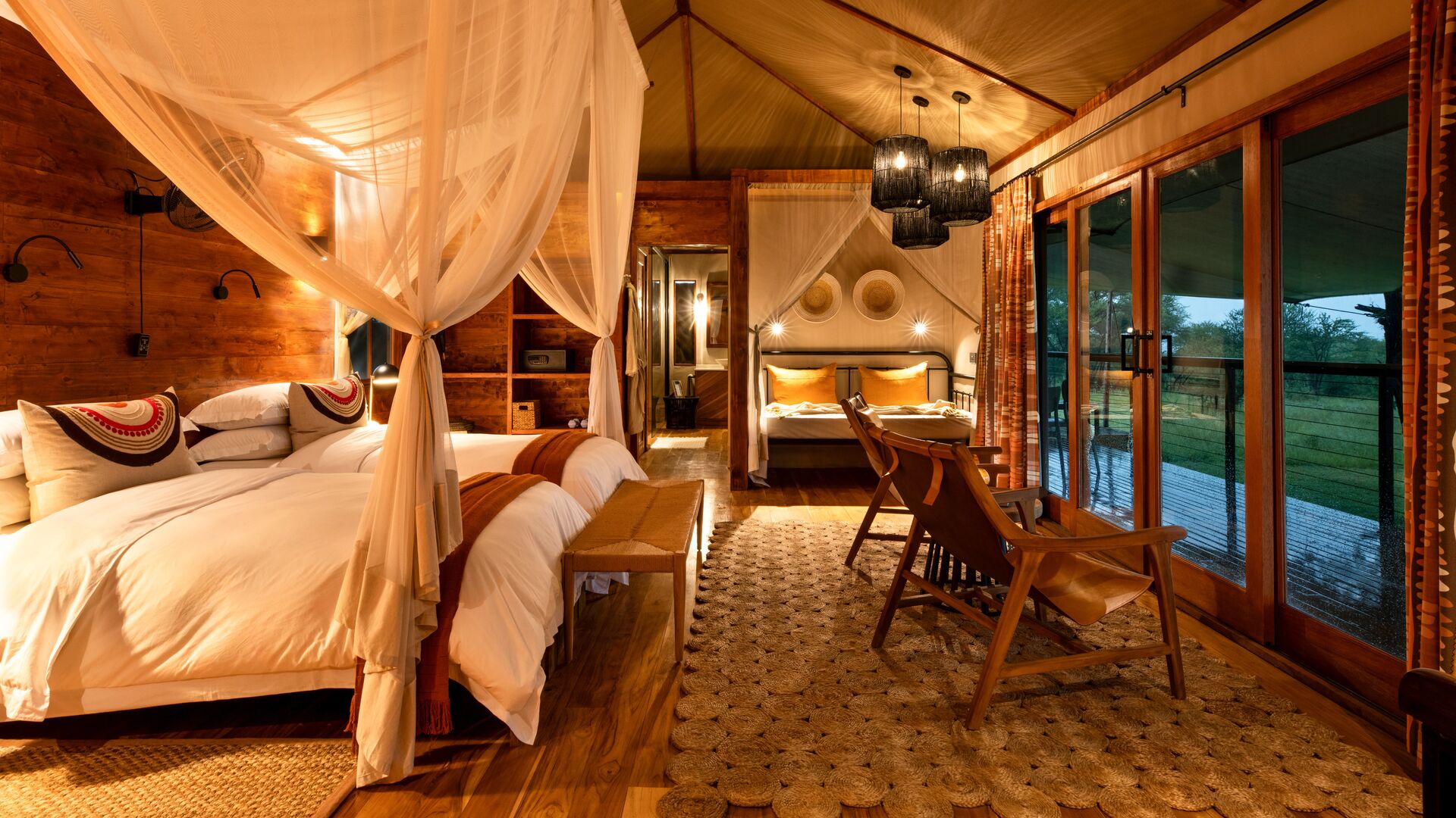 Interior of a family room at Sayari Retreat featuring two canopy beds, warm wooden interiors, soft ambient lighting, woven rugs, lounge chairs, and large glass doors opening onto a private deck overlooking the surrounding landscape.