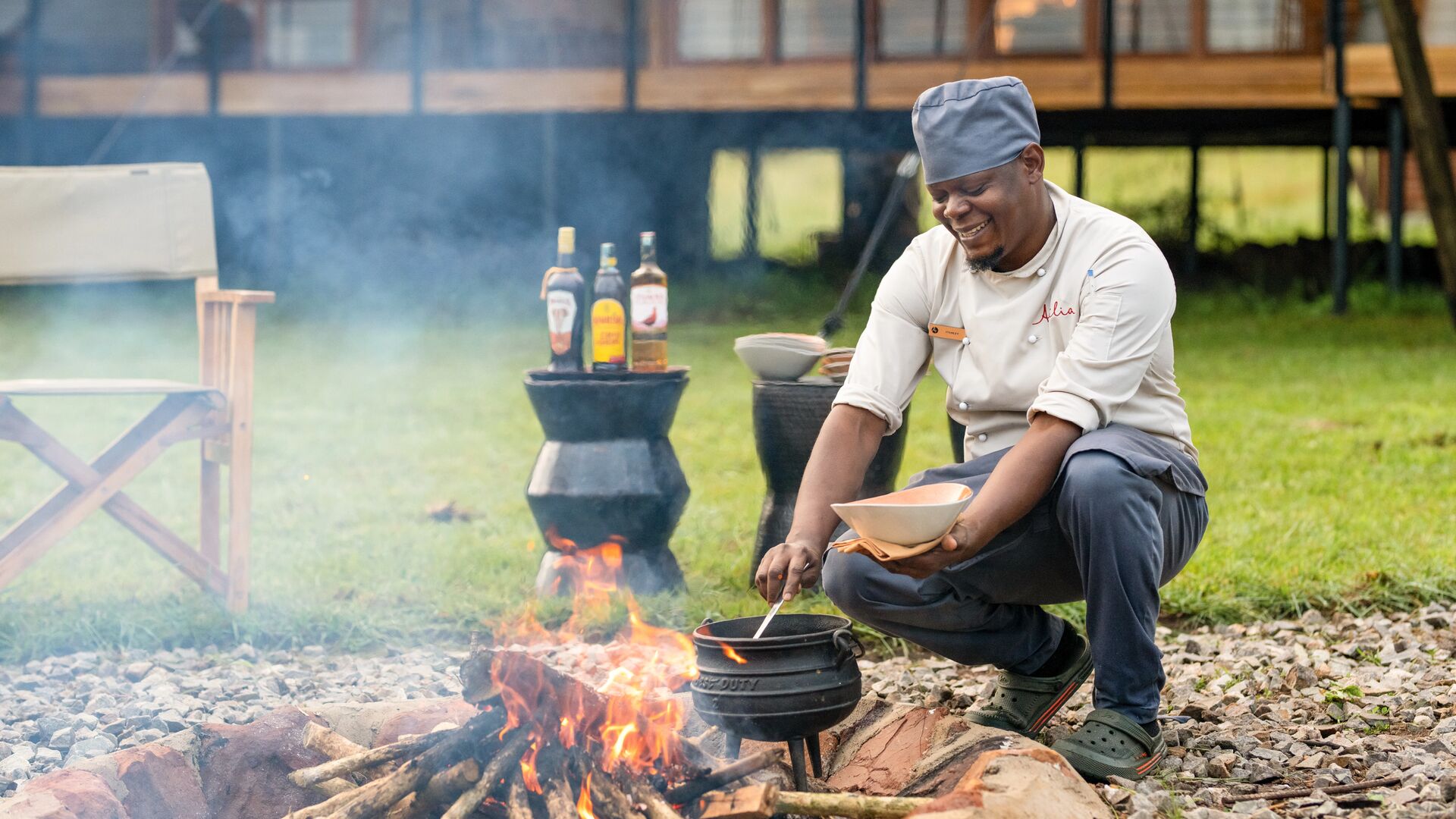 A chef from Sayari Retreat crouches beside an open campfire, stirring food in a cast-iron pot set over flames, with smoke rising, cooking utensils and bottles nearby, and the lodge buildings visible in the background on a grassy lawn.