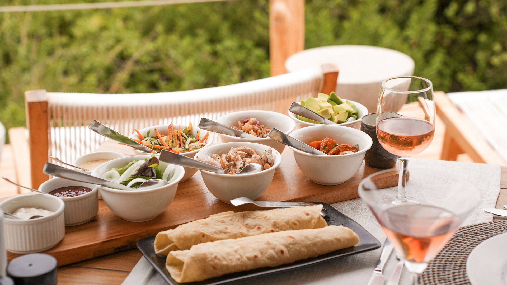 Outdoor lunch spread on a wooden table featuring wraps, bowls of salads, grains, chicken, avocado, sauces, and glasses of rosé wine, set on a deck overlooking lush green forest.