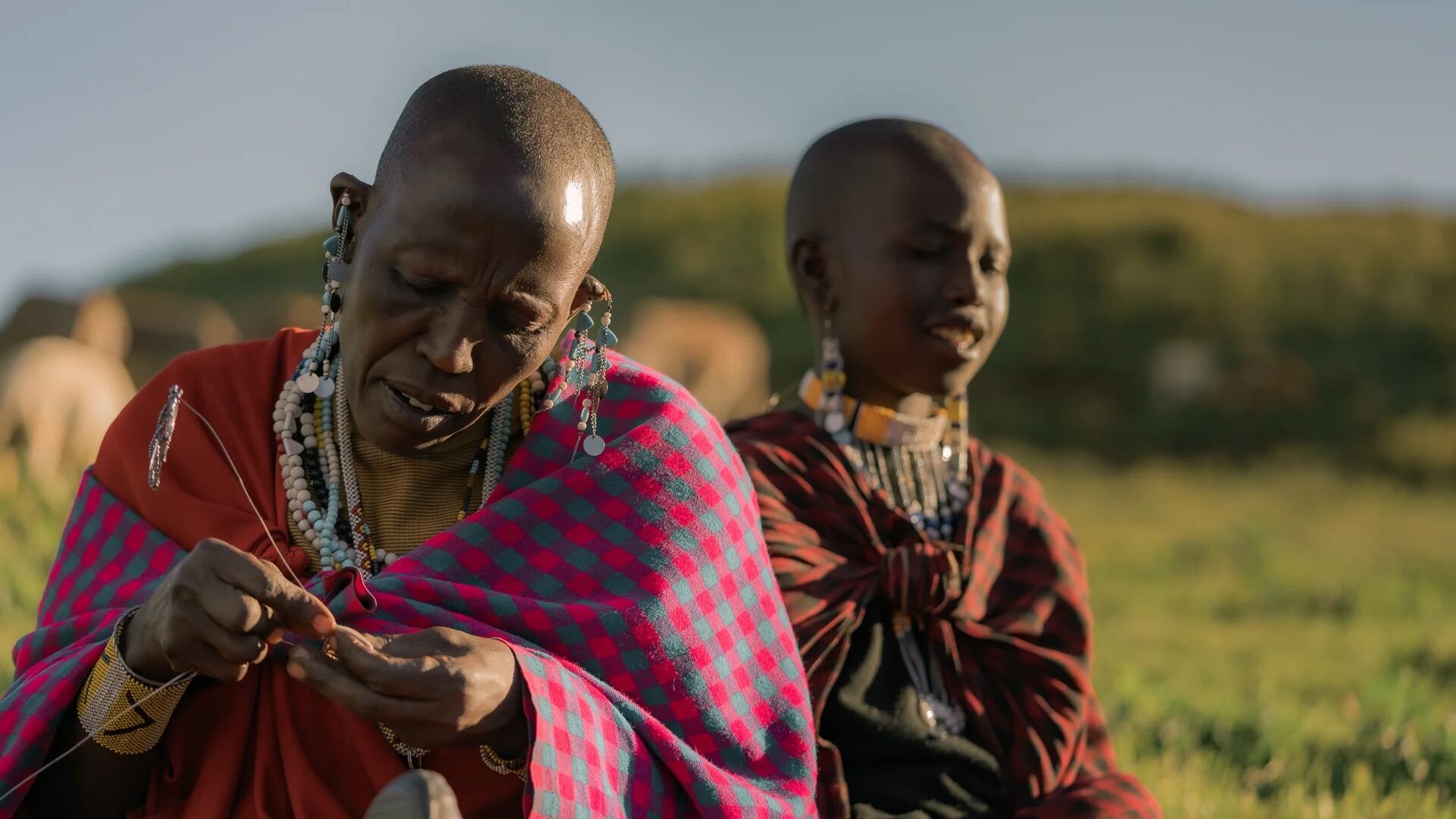 Two Maasai women seated outdoors on open grassland, wearing traditional red and patterned shukas and beaded jewelry, carefully handcrafting beadwork together in warm afternoon light.