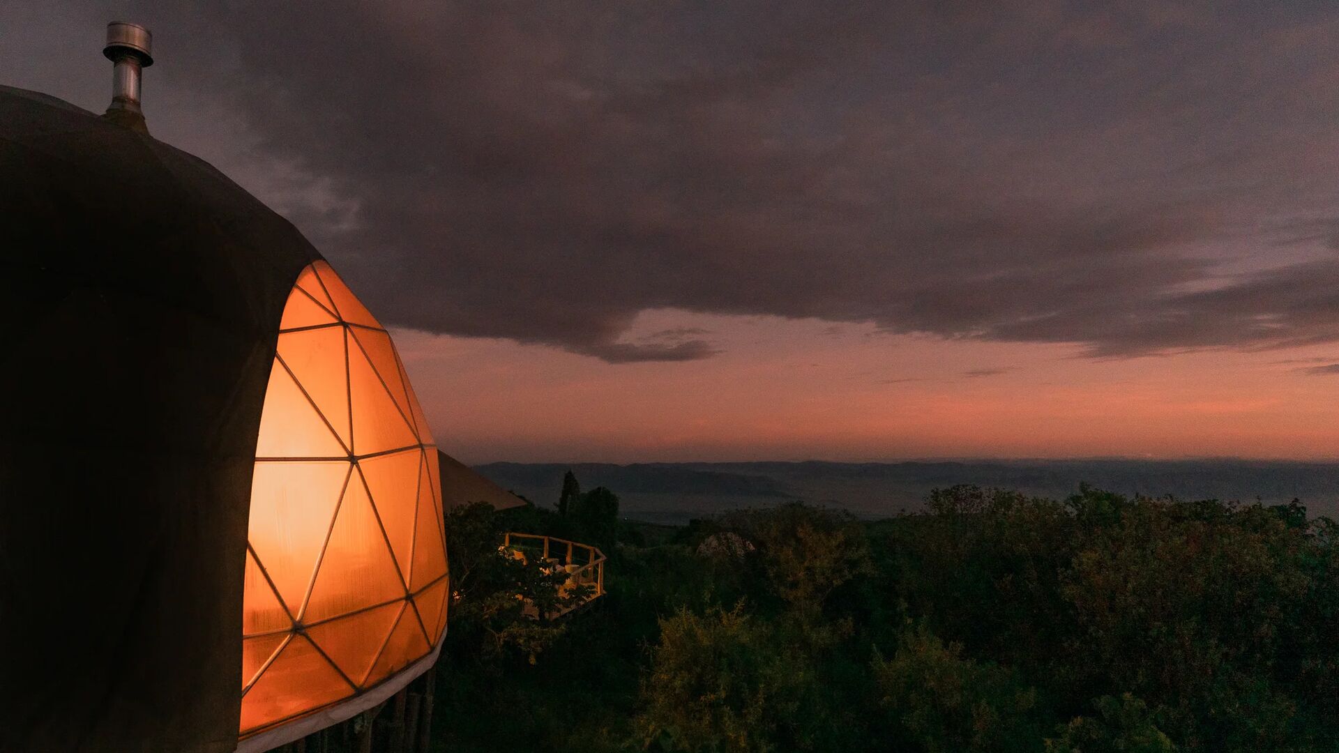 A softly lit geodesic dome at The Highlands Camp at dusk, glowing warmly against dark clouds and a fading pink horizon, overlooking forested slopes and distant highlands.