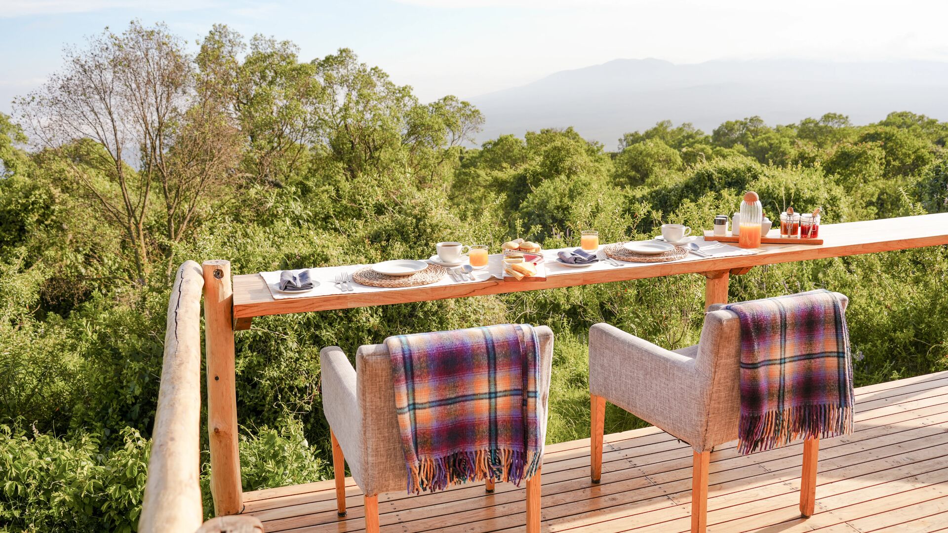 Outdoor dining setup at The Highlands on a wooden deck with a long counter table set for breakfast, cushioned chairs draped with blankets, and views over dense green forest toward distant mountains under soft morning light.