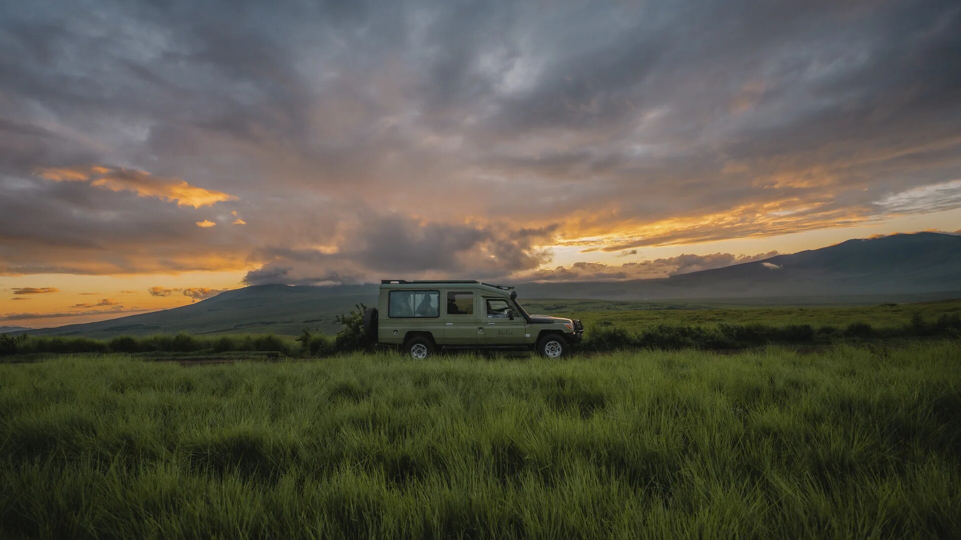 An Asilia safari vehicle parked on a grassy plain at sunset, with dramatic clouds glowing orange and purple over rolling highland hills and distant mountains.