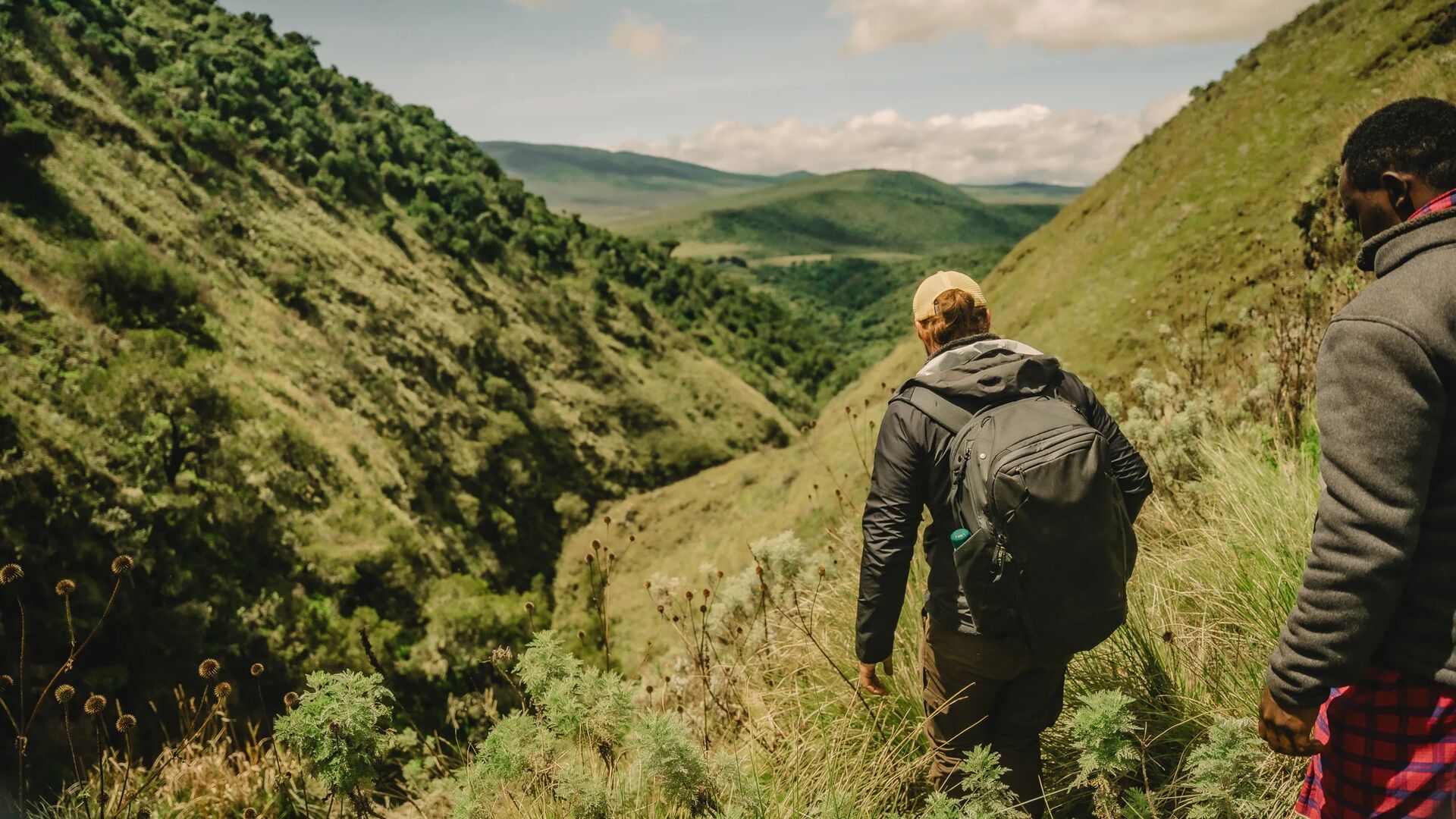 Two people hiking along a narrow grassy trail through a steep green valley, carrying backpacks and overlooking rolling hills and layered highland landscapes under a bright sky.