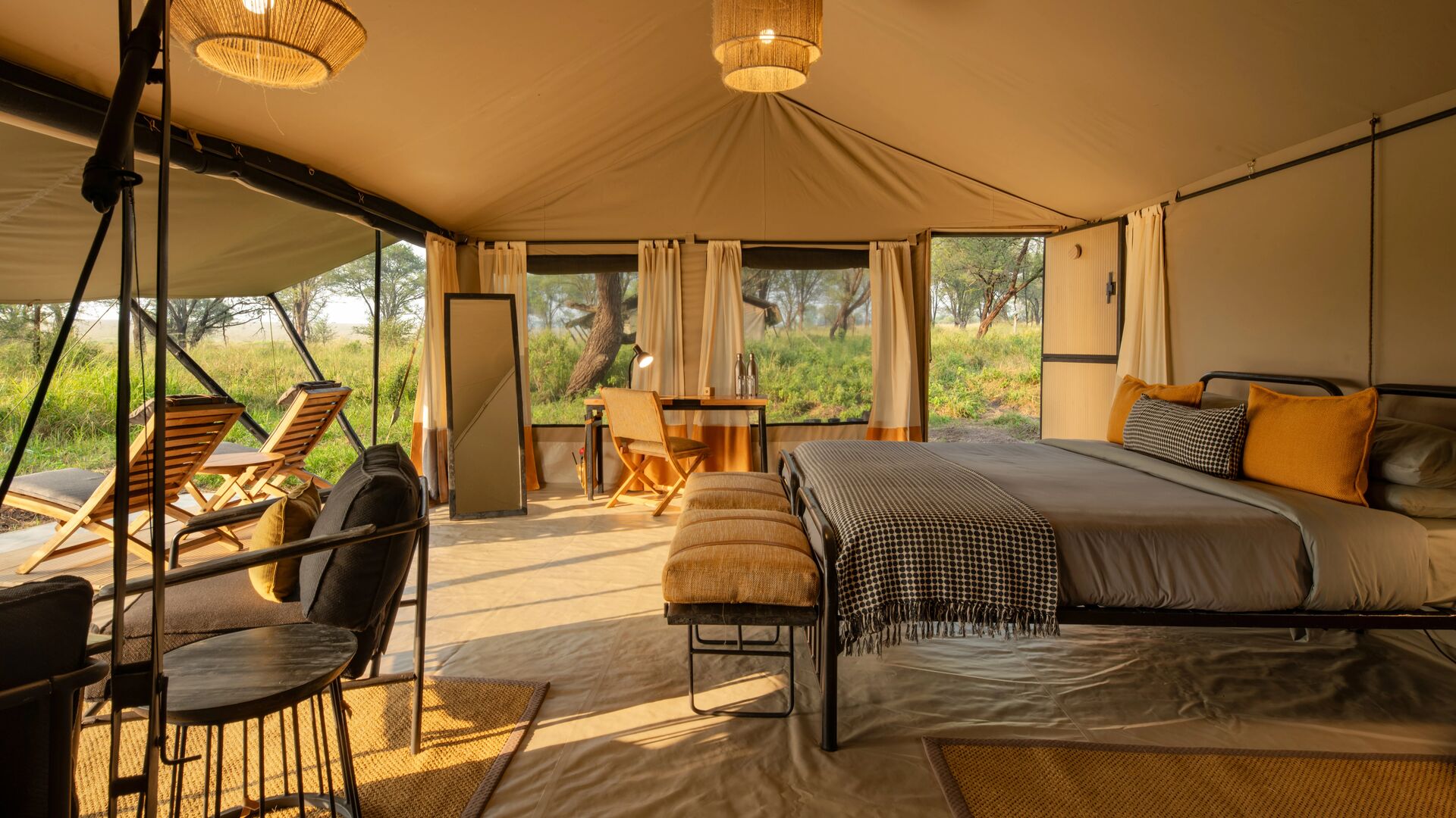Interior of a safari bedroom at Ubuntu Migration Camp with a double bed, woven lighting, seating area, writing desk, and open canvas walls revealing green woodland and outdoor loungers.