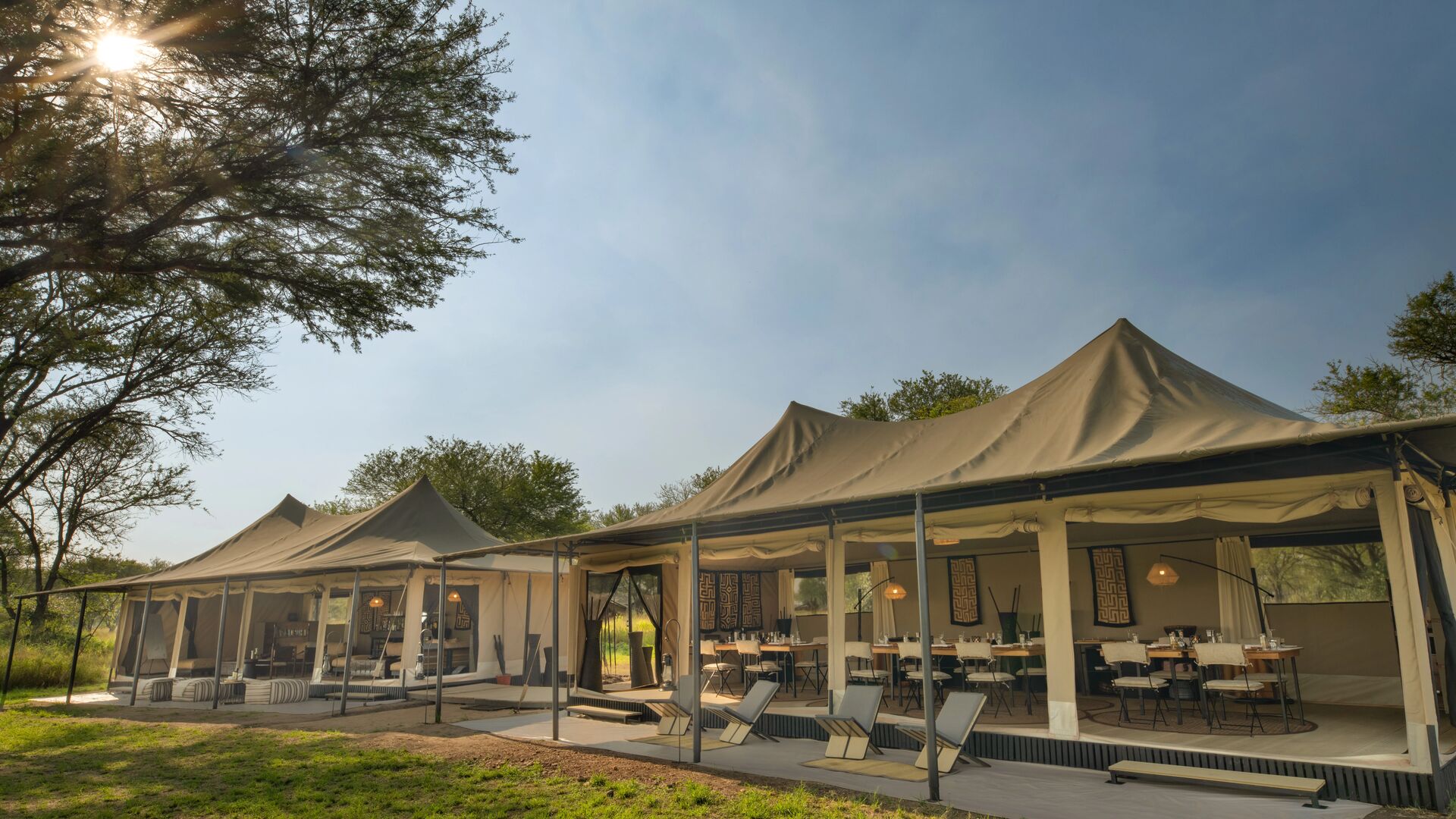 ~Ubuntu Migration Camp's open-sided communal safari tents with dining and lounge areas, canvas roofs and furnishings, set beneath trees with sunlight filtering through onto the grassy clearing.