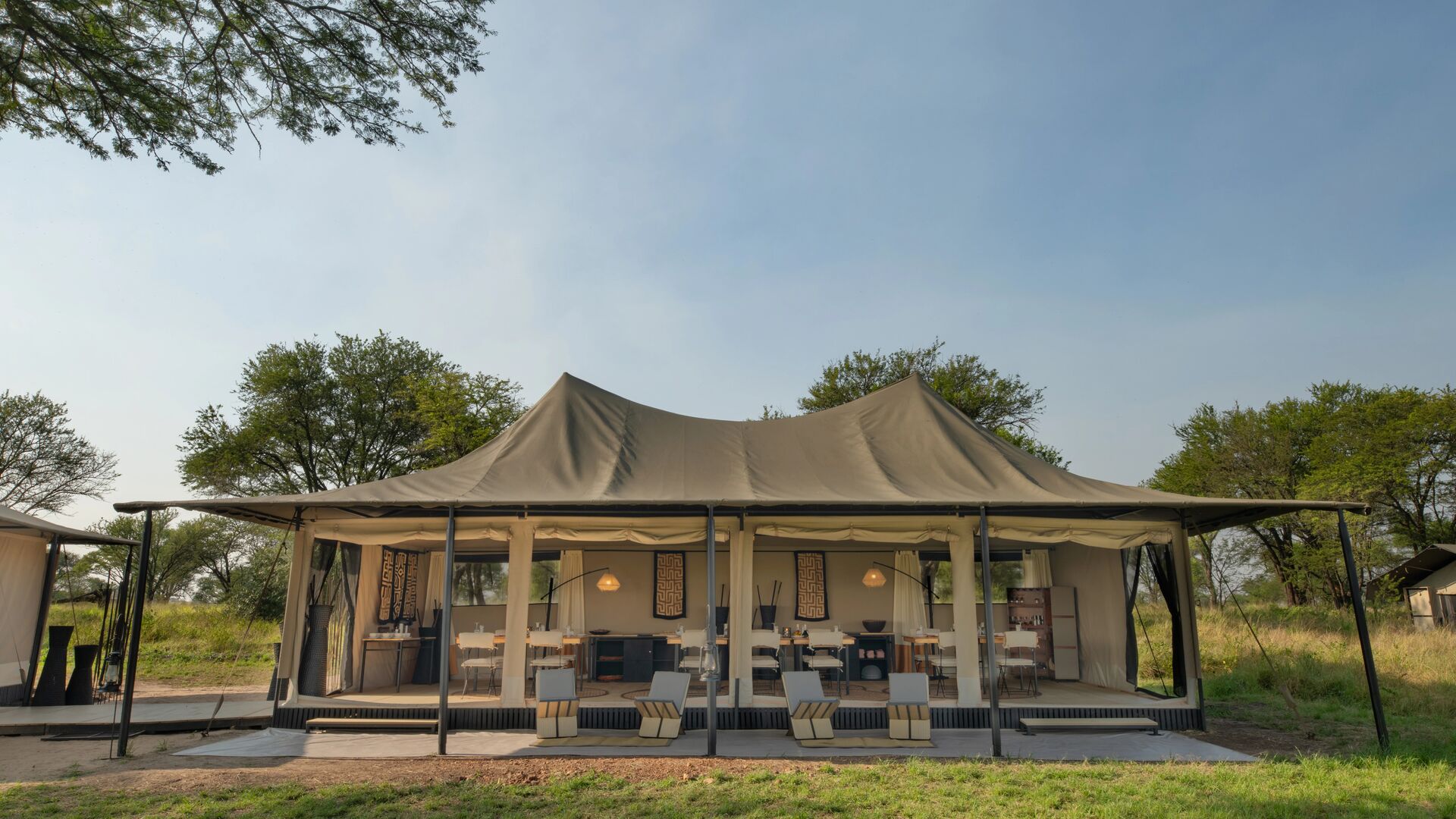 Ubuntu Migration Camp's spacious open-sided safari dining tent with neatly arranged tables and chairs, canvas walls, and modern furnishings, set among trees and grassland under a clear sky.