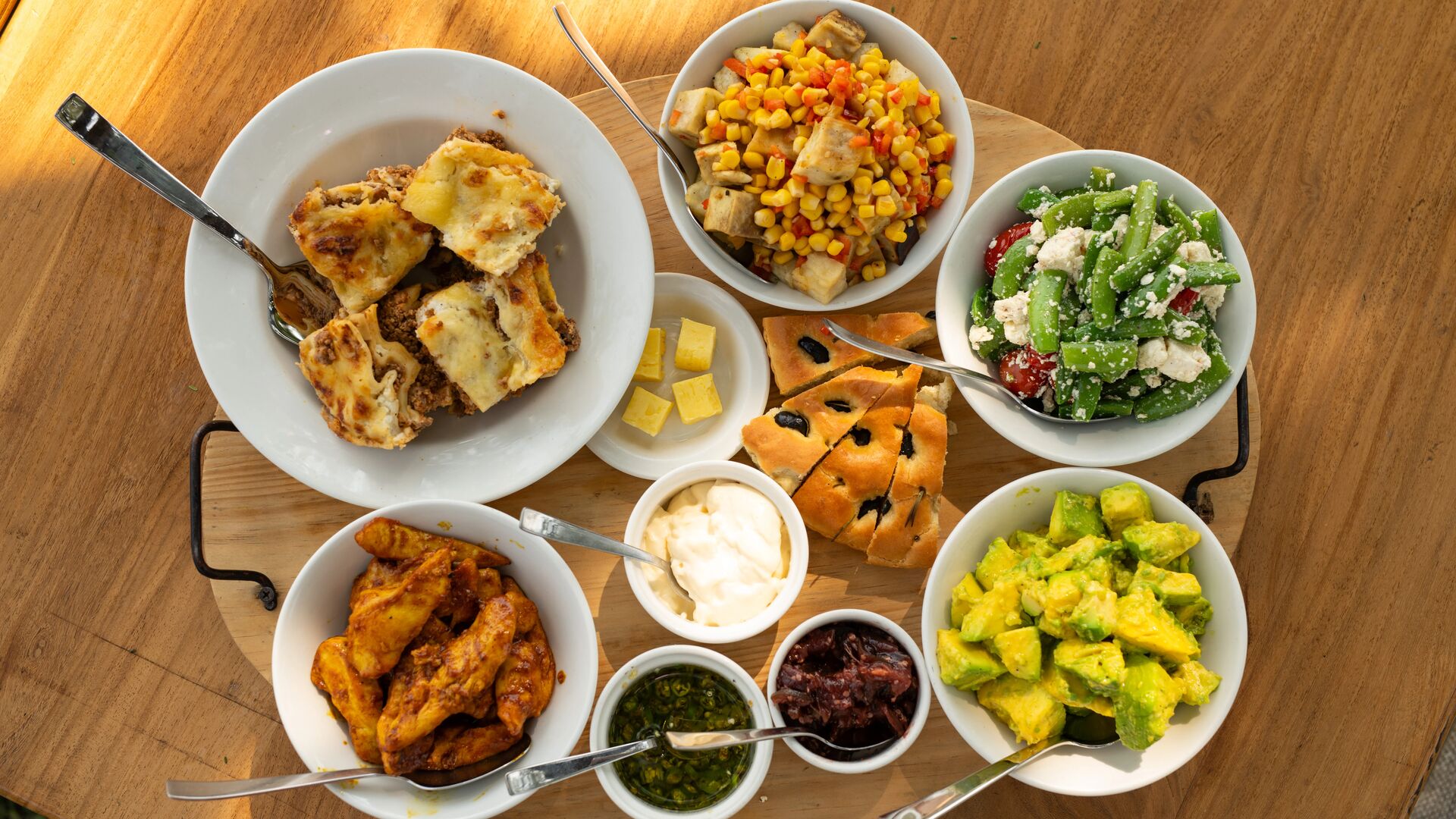 Overhead view of a safari lunch spread on a wooden table, featuring lasagne, roasted chicken, avocado salad, green bean and feta salad, corn and vegetable salad, focaccia bread, and assorted sauces in white bowls.