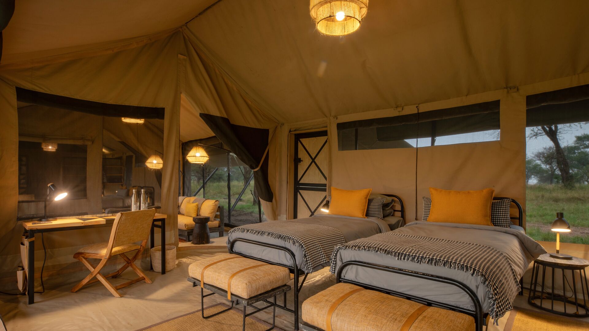 Interior of a family suite featuring two single beds with yellow cushions, soft ambient lighting, a writing desk, and canvas walls opening to views of surrounding grassland.
