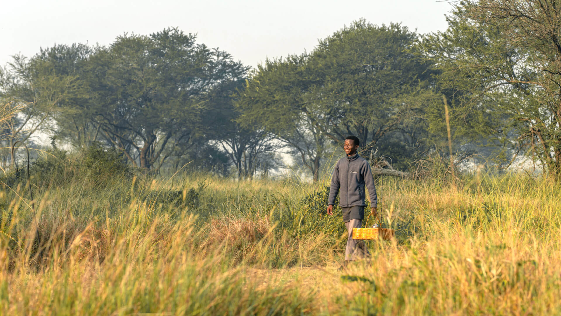 Smiling Asilia staff member walks through long grass carrying a basket
