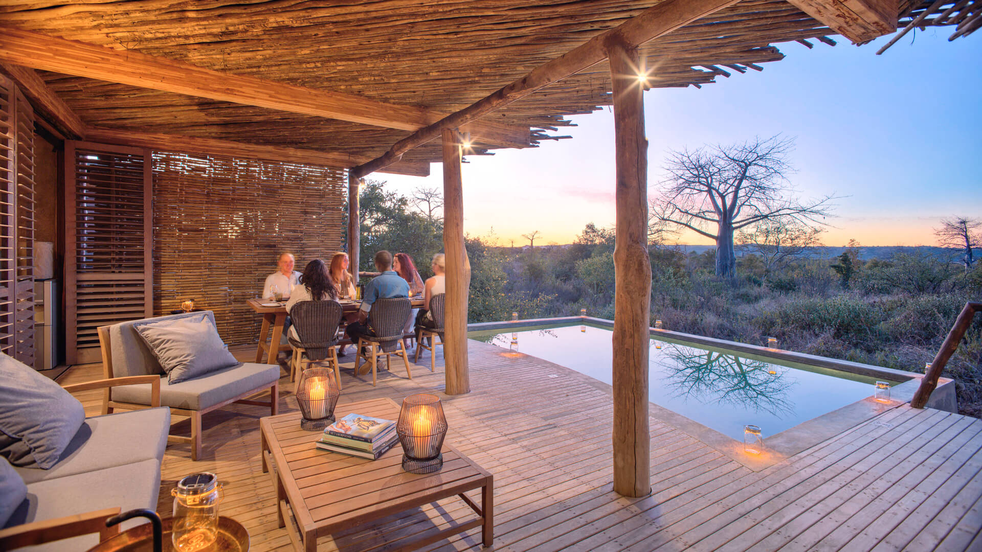 Jabali Private House, Ruaha National Park, guests having dinner by the pool lit up with lanterns at sunset, deck loungers, table with books, and candles