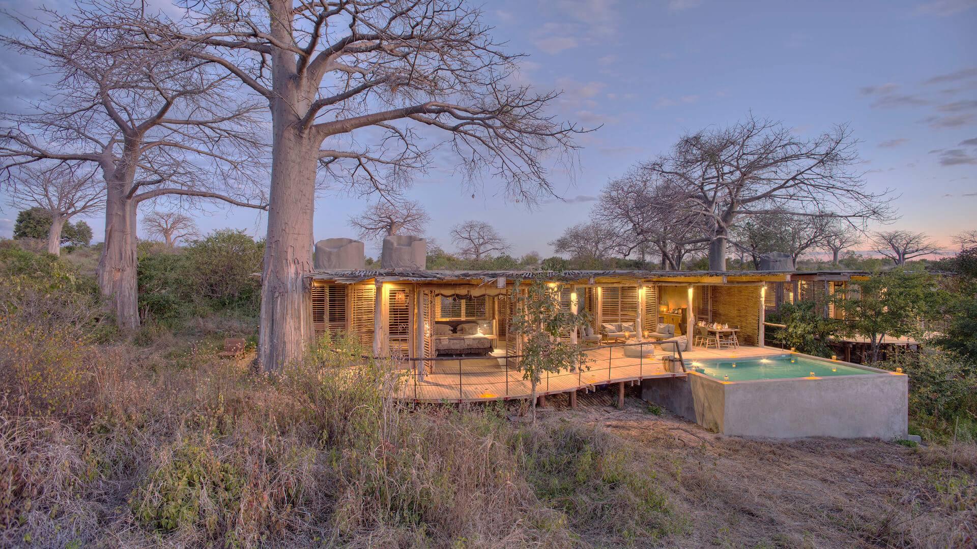 Jabali Private House, Ruaha National Park, view of the outside at sunset with pool and lanterns lighting up the deck with tables and chairs and open shutters to the room