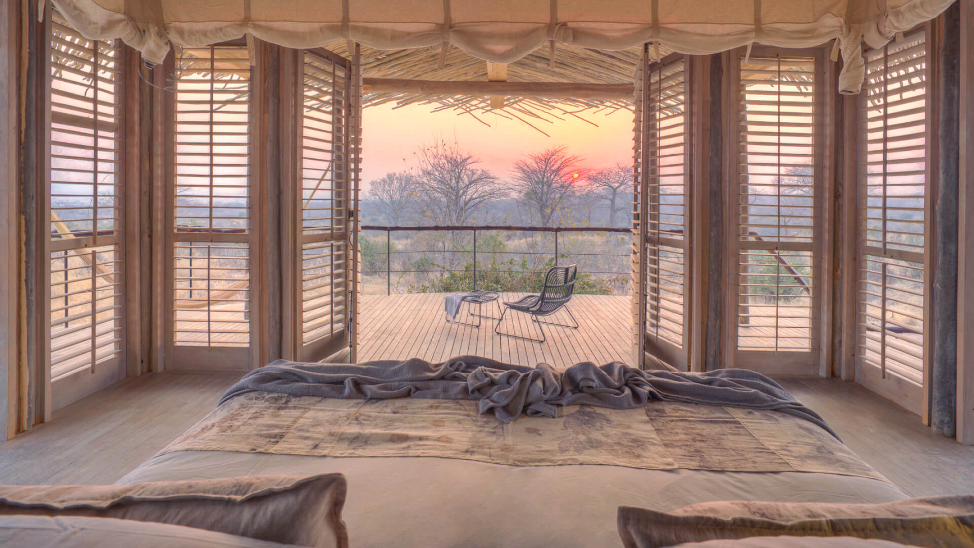 Jabali Ridge, Ruaha National Park, double bed with pillows and throw blanket, open shutters leading out to the deck with table and chair