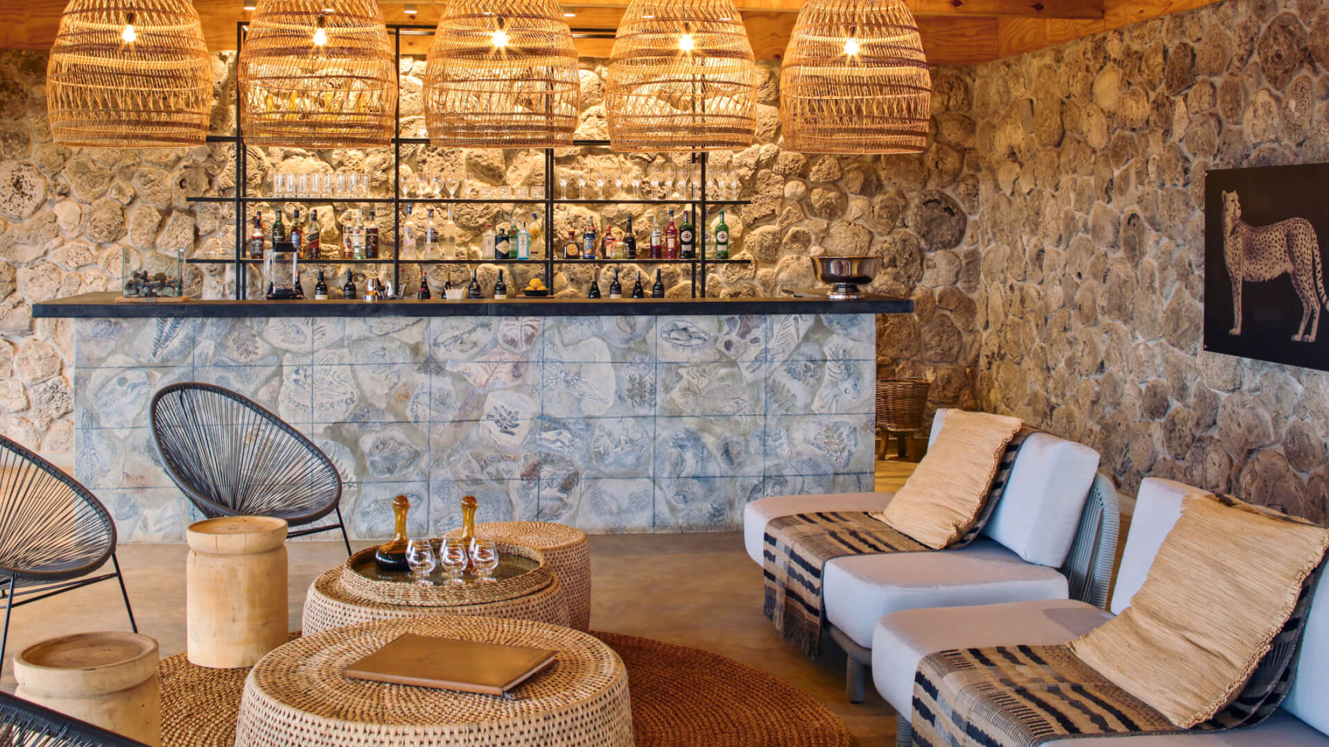 Namiri Plains, Serengeti National Park, bar at the main area, couches and wicker tables and chairs with shanga glass ornaments, hanging lights and cheetah infographic on the wall