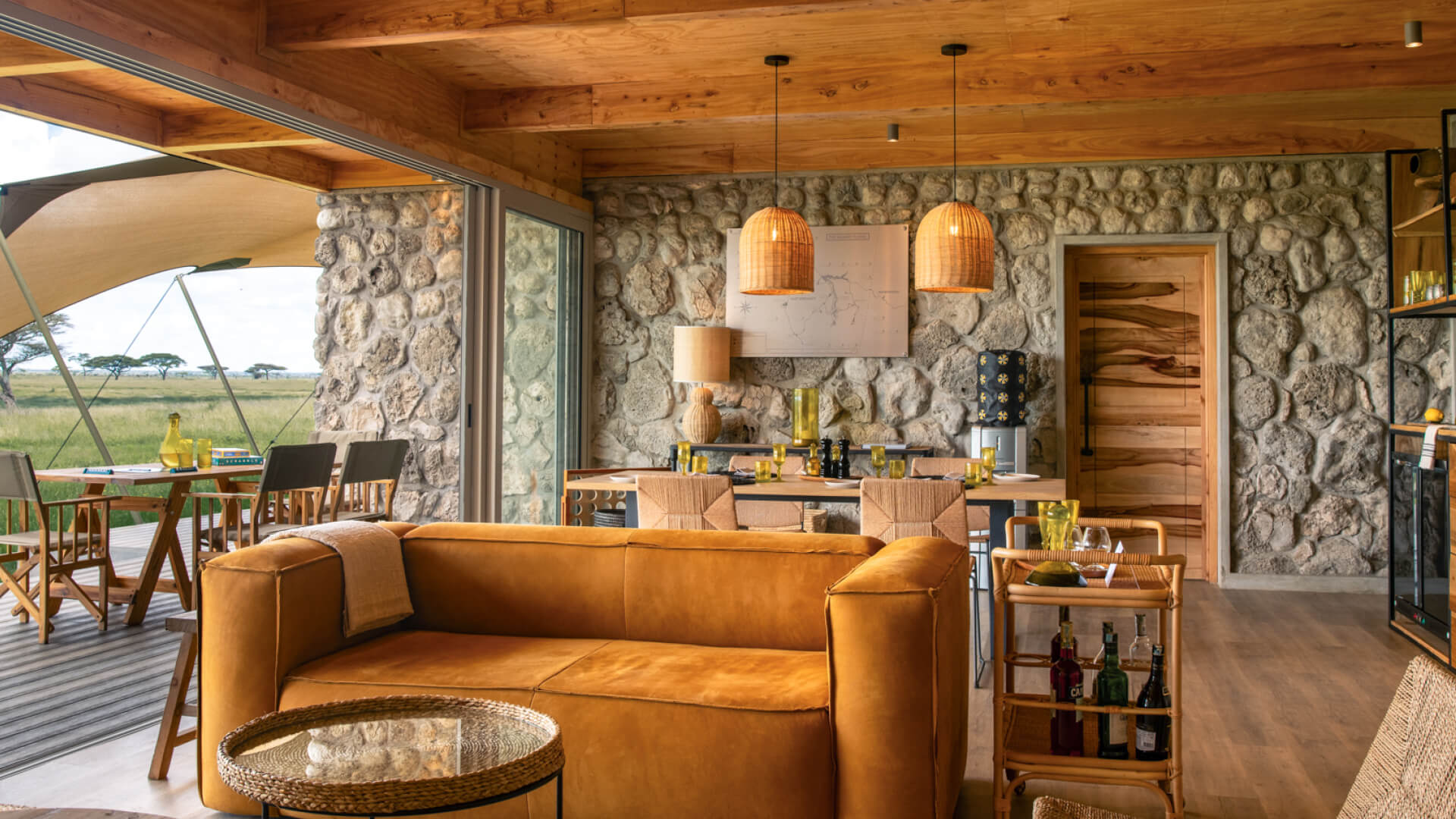 Namiri Plains Retreat, Serengeti National Park, lounge area interior, couch and table, tray of drinks and dining table setup for lunch, hanging lights and infographic on the wall