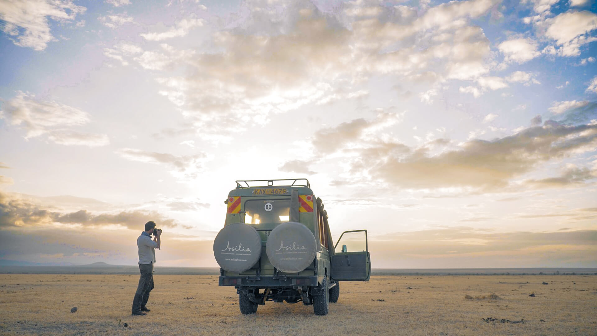 A photographer stands next to a safari vehicle and photographs the sunset
