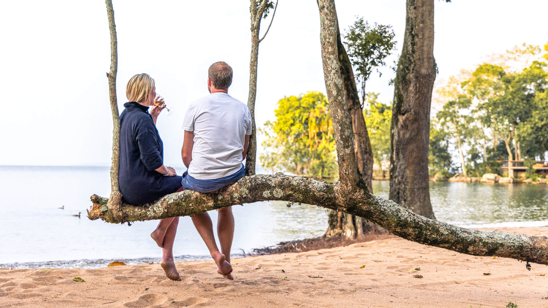 A couple enjoy a sundowner whilst sat on a branch on the beach at Rubondo Island Camp in Tanzania - Asilia Africa