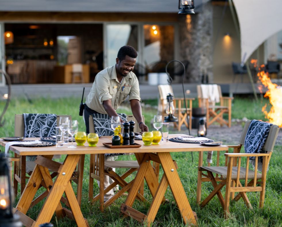 Namiri Plains Retreat, Serengeti National Park, butler setting up dinner table by the campfire, chairs with shukas and table with condiments and shanga glass candles and wine glasses