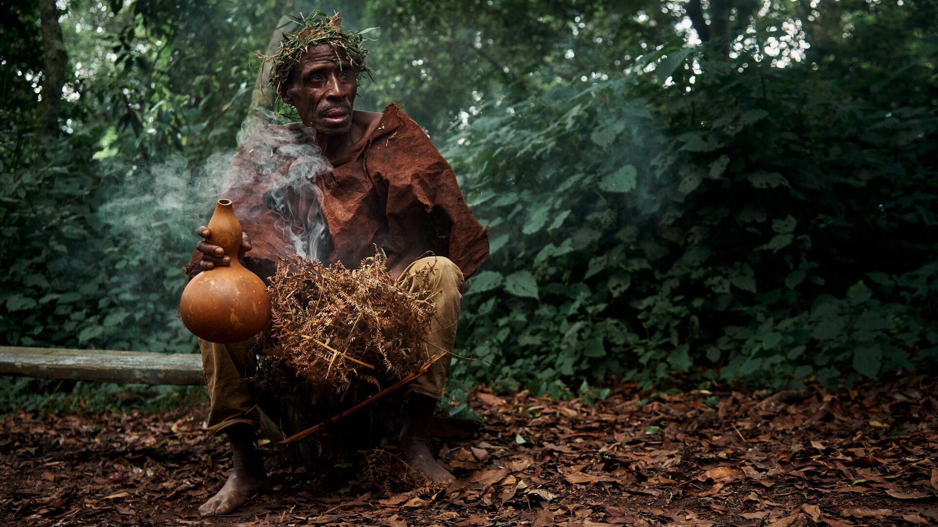 An elderly Batwa man holding a gourd and herbs, seated in a forest with smoke rising around him