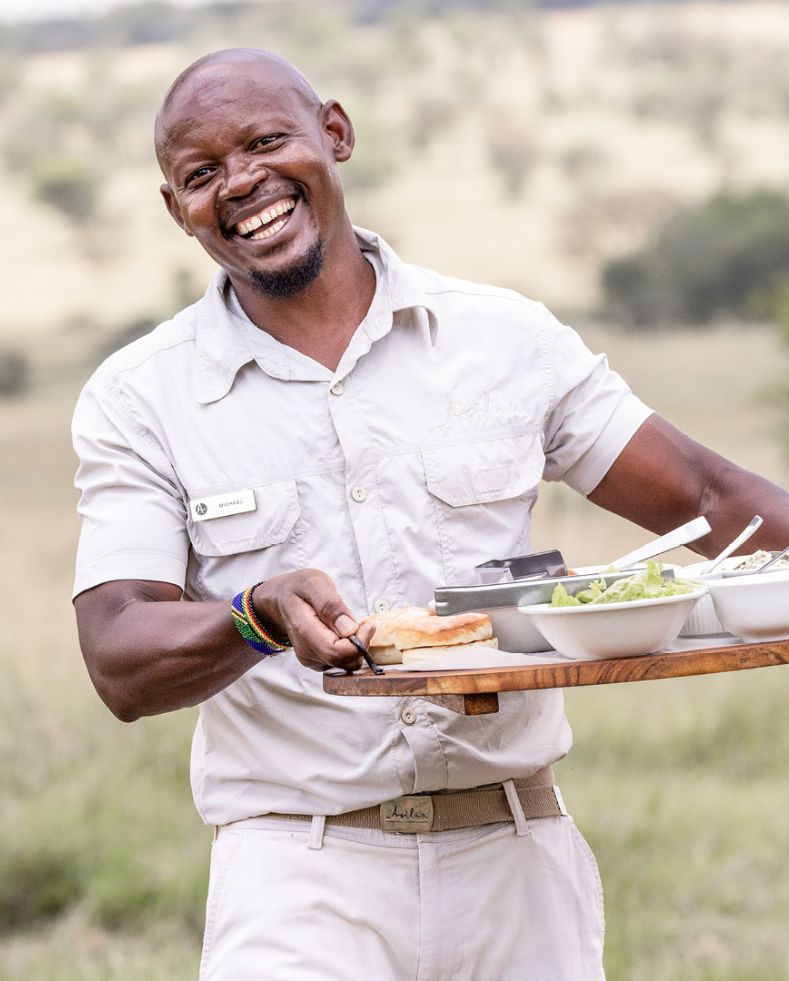 male waiter walking towards the camera smiling, serving guests lunch at olakira migration camp, tanzania, asilia africa