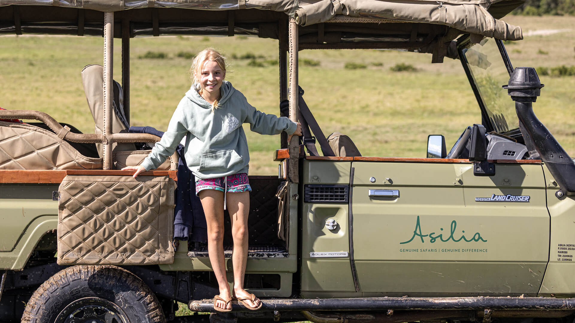 Young girl standing on the Asilia vehicle wearing shorts, flip flops and a hoodie
