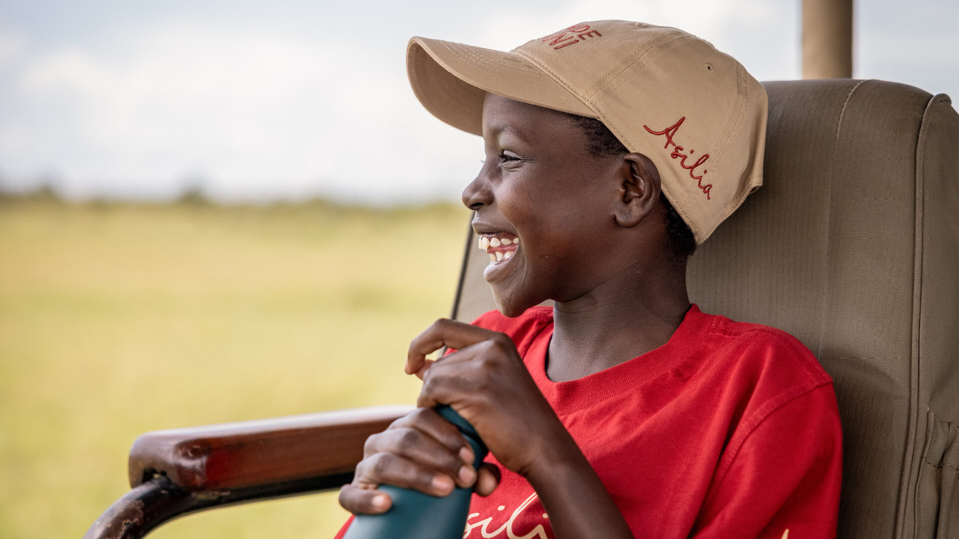 Young boy in a red t-shirt holding Asilia water bottle wearing Asilia cap smiling