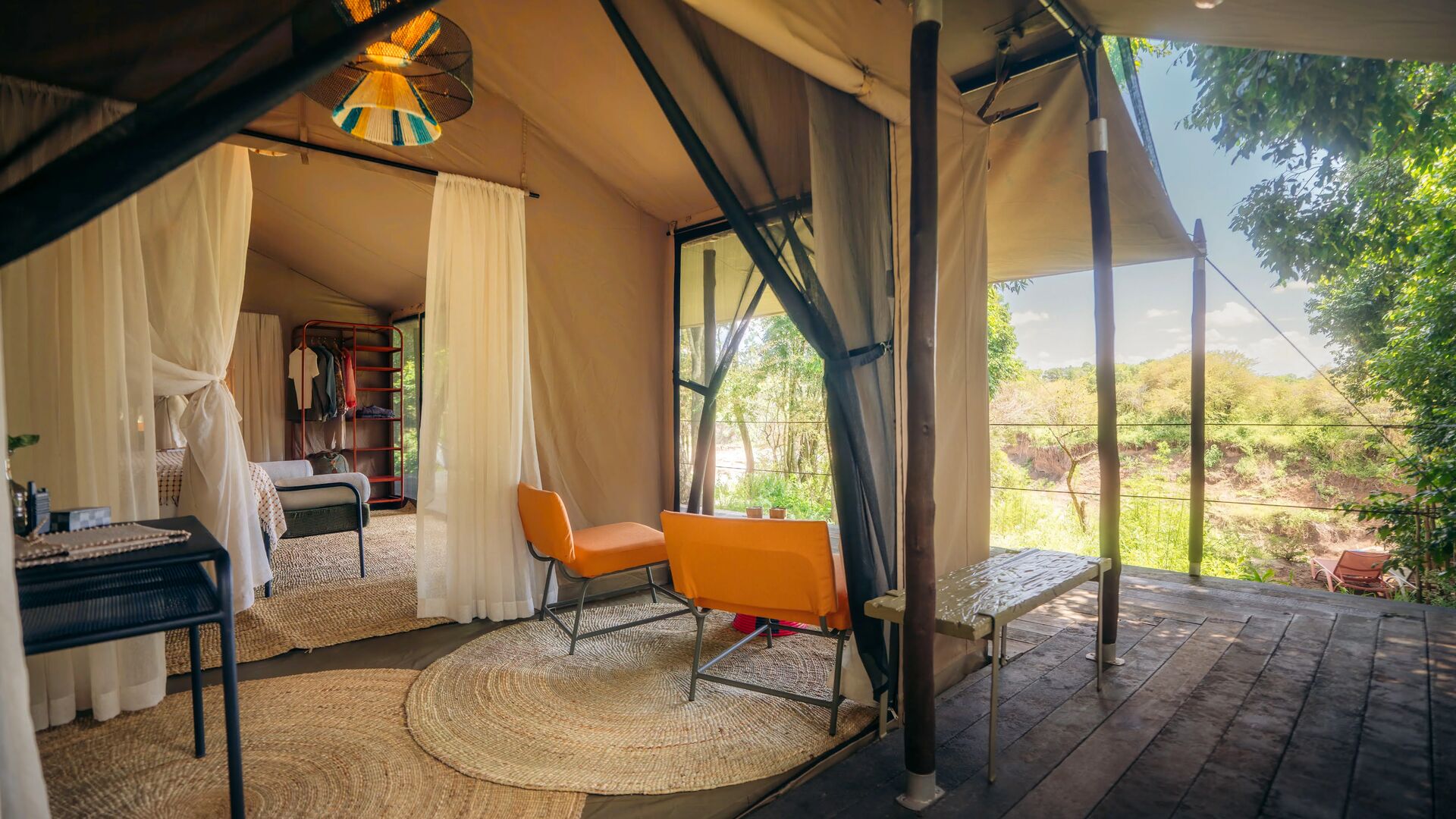 Spacious luxury safari tent at Emboo with bed, seating area, woven rugs, open canvas walls, and a private wooden deck overlooking a green riverbank landscape.