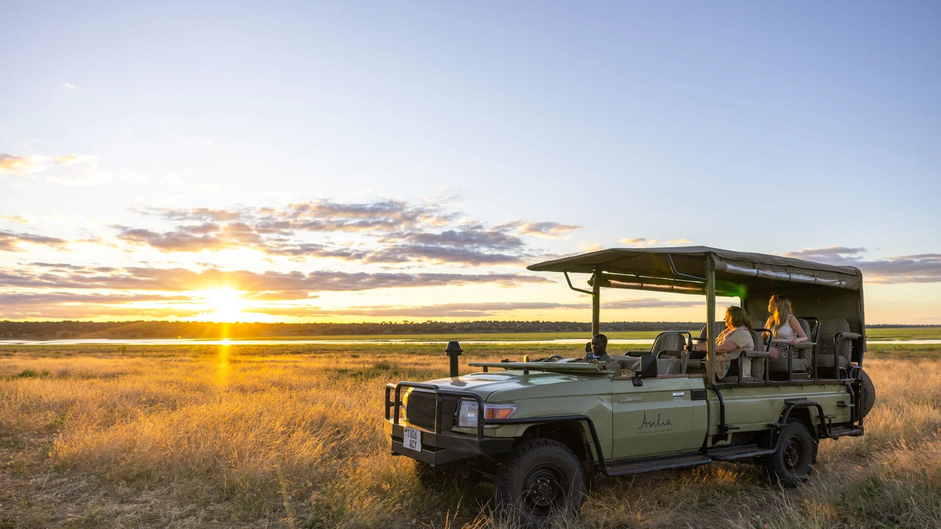 An open safari vehicle with guests seated inside pauses in tall golden grass at sunset, overlooking open plains and distant water as warm light spreads across the sky. Oliver's Camp, Tarangire, Asilia Africa